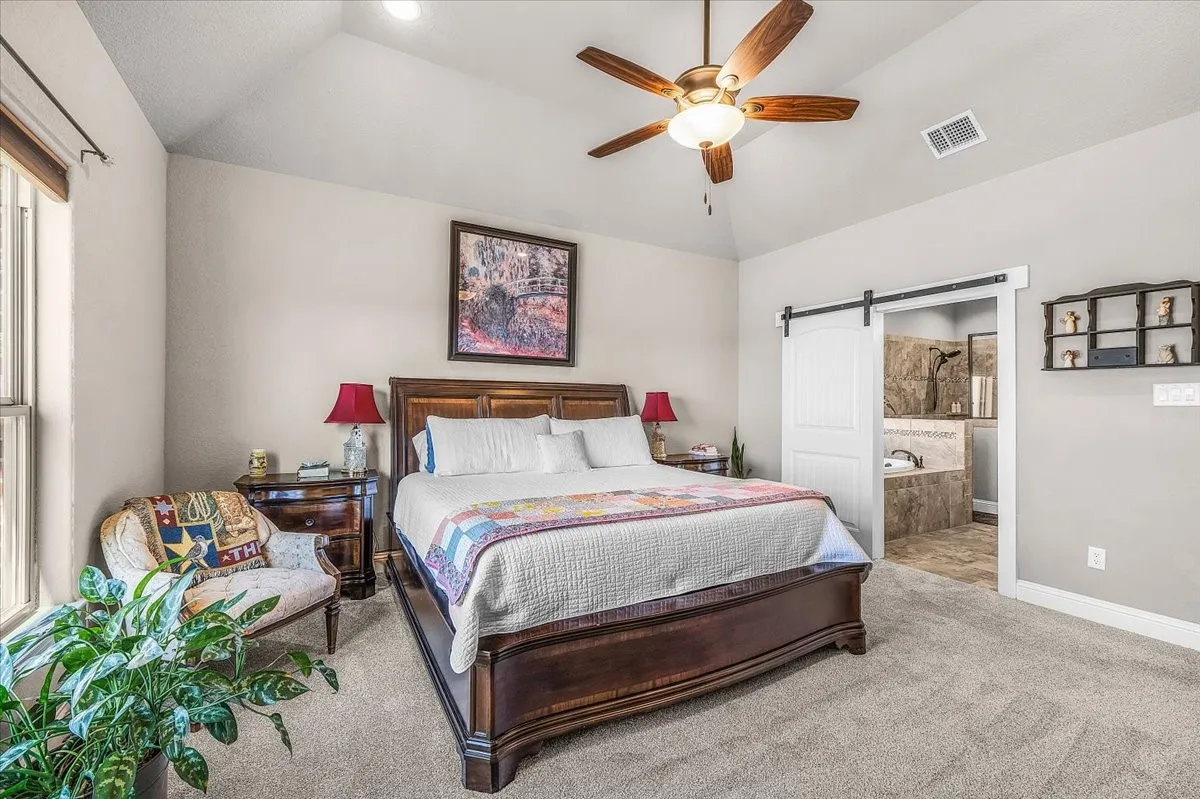 Primary Bedroom: Spacious primary bedroom features a vaulted ceiling, ceiling fan, plush carpeting, and direct access to the en-suite bath through a sliding barn-style door.
