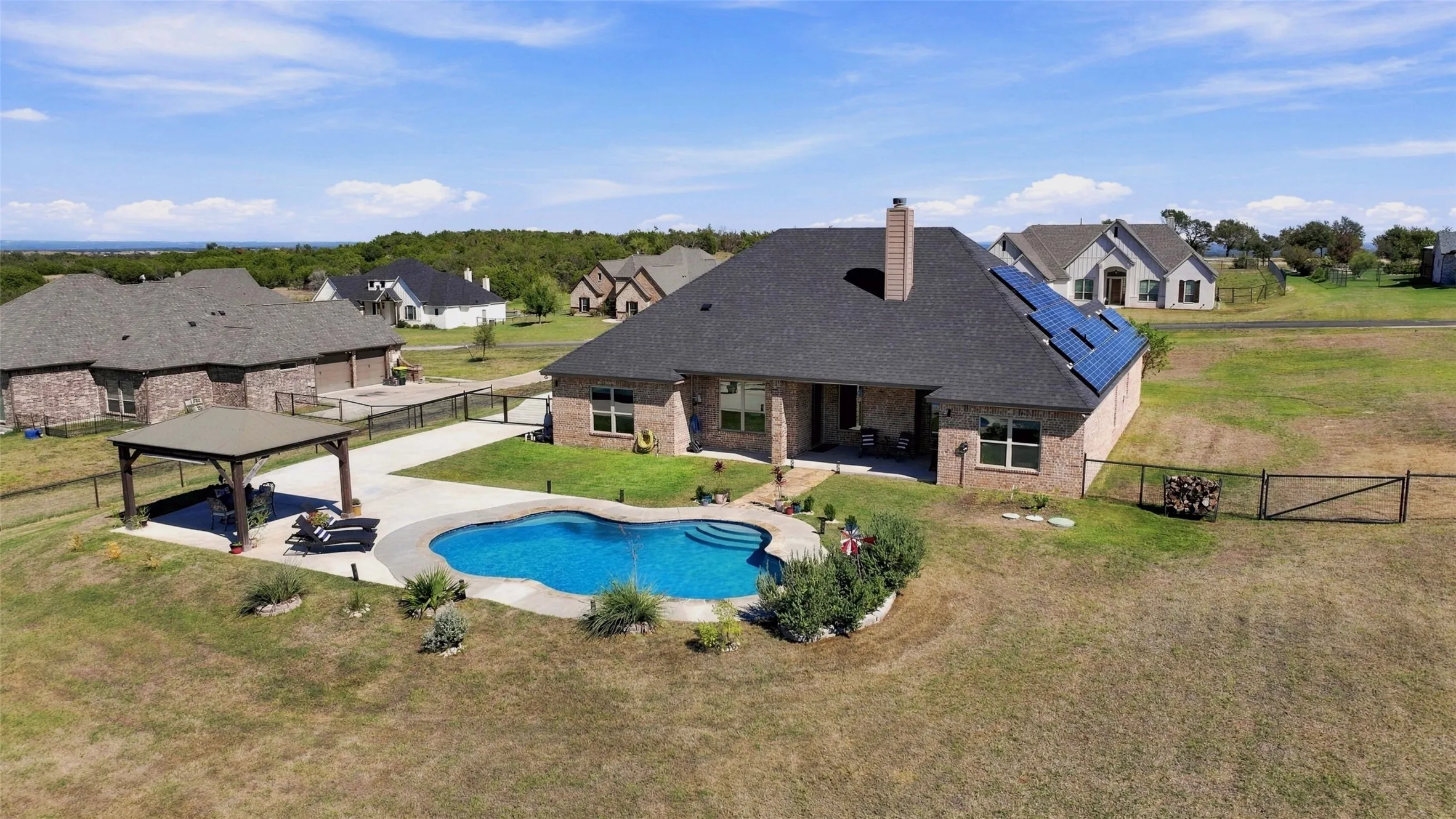 Aerial view showcasing the sparkling in-ground pool, covered outdoor pavilion, expansive patio, and spacious fenced backyard, perfect for entertaining and outdoor enjoyment.