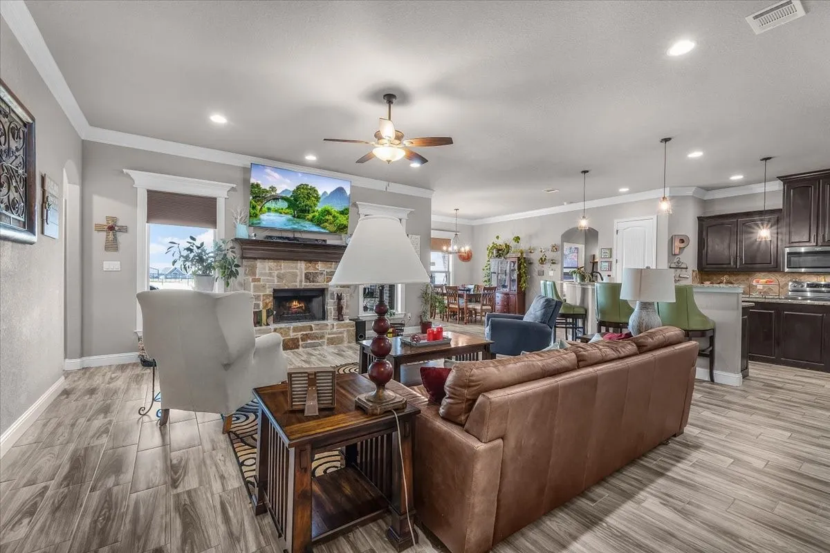 Living Room / Open-Concept View: Spacious living area showcases a stone fireplace, crown molding, abundant recessed lighting, and an open connection to the kitchen and dining spaces.
