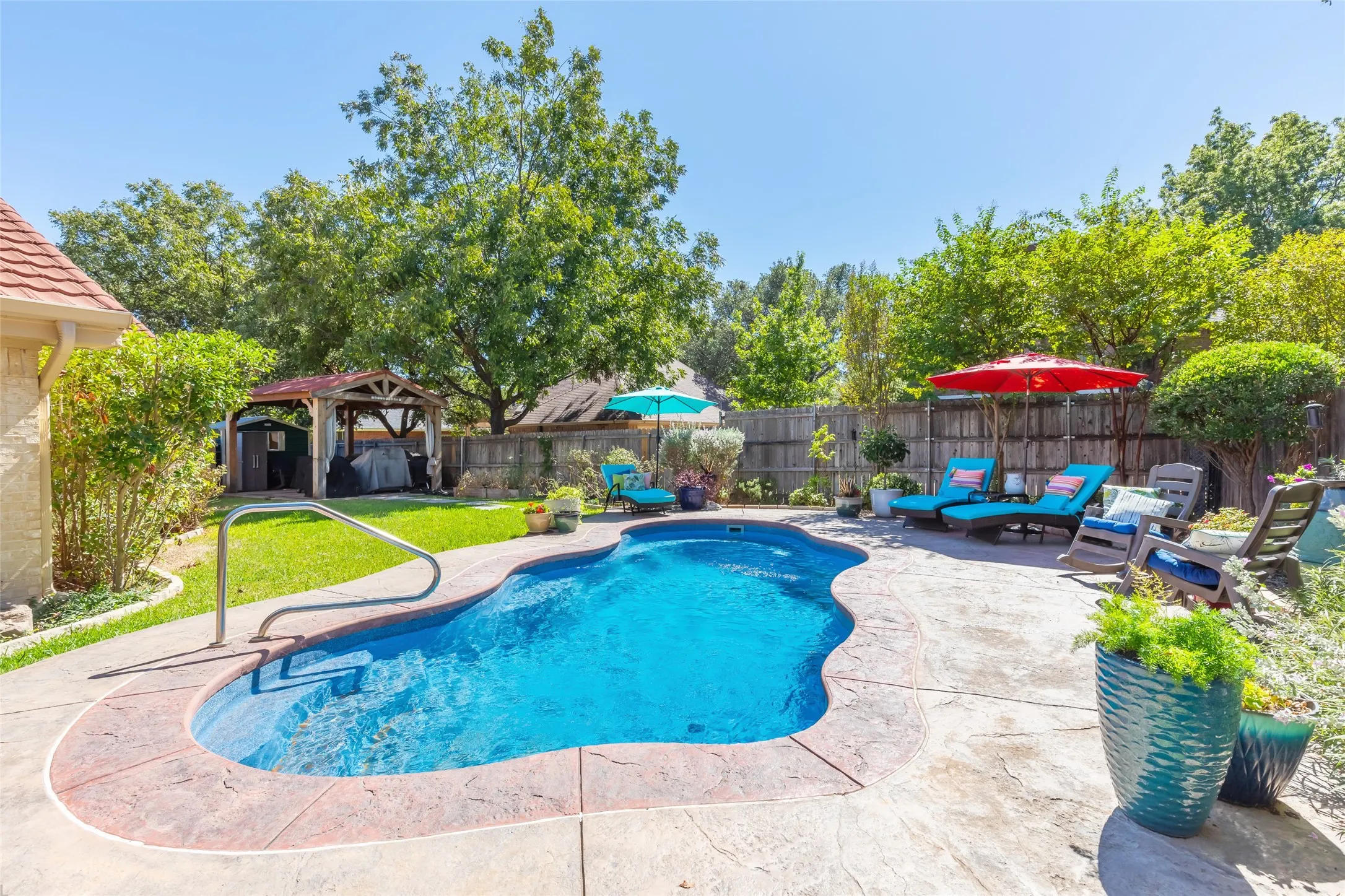 Freeform swimming pool with a surrounding stamped concrete deck, a built-in pool handrail, and a backyard gazebo