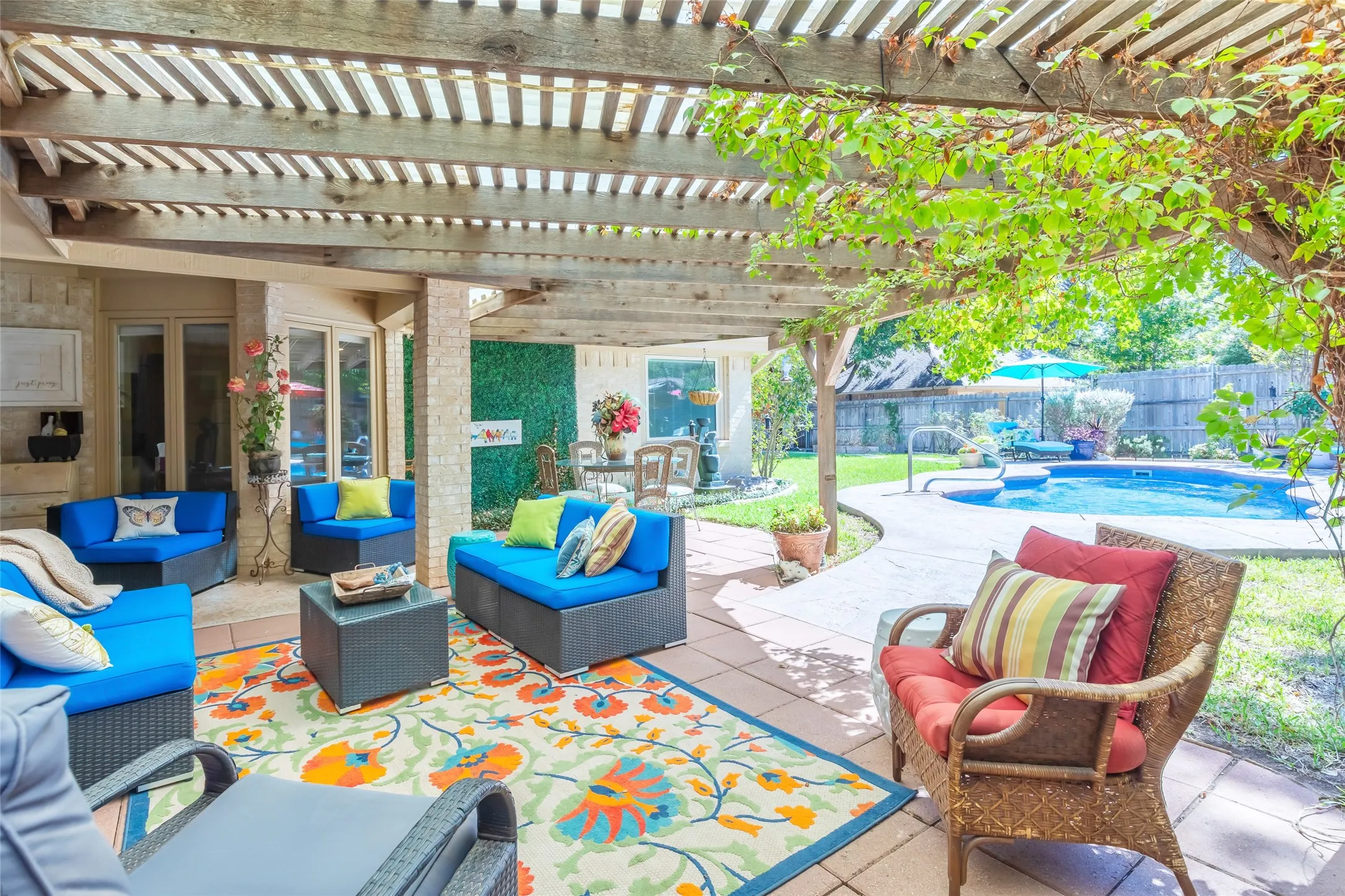 Covered lounge area featuring a slatted pergola with climbing vines, vibrant blue upholstered seating, patterned tile flooring, and direct access to resort-style backyard with in-ground swimming pool and mature landscaping