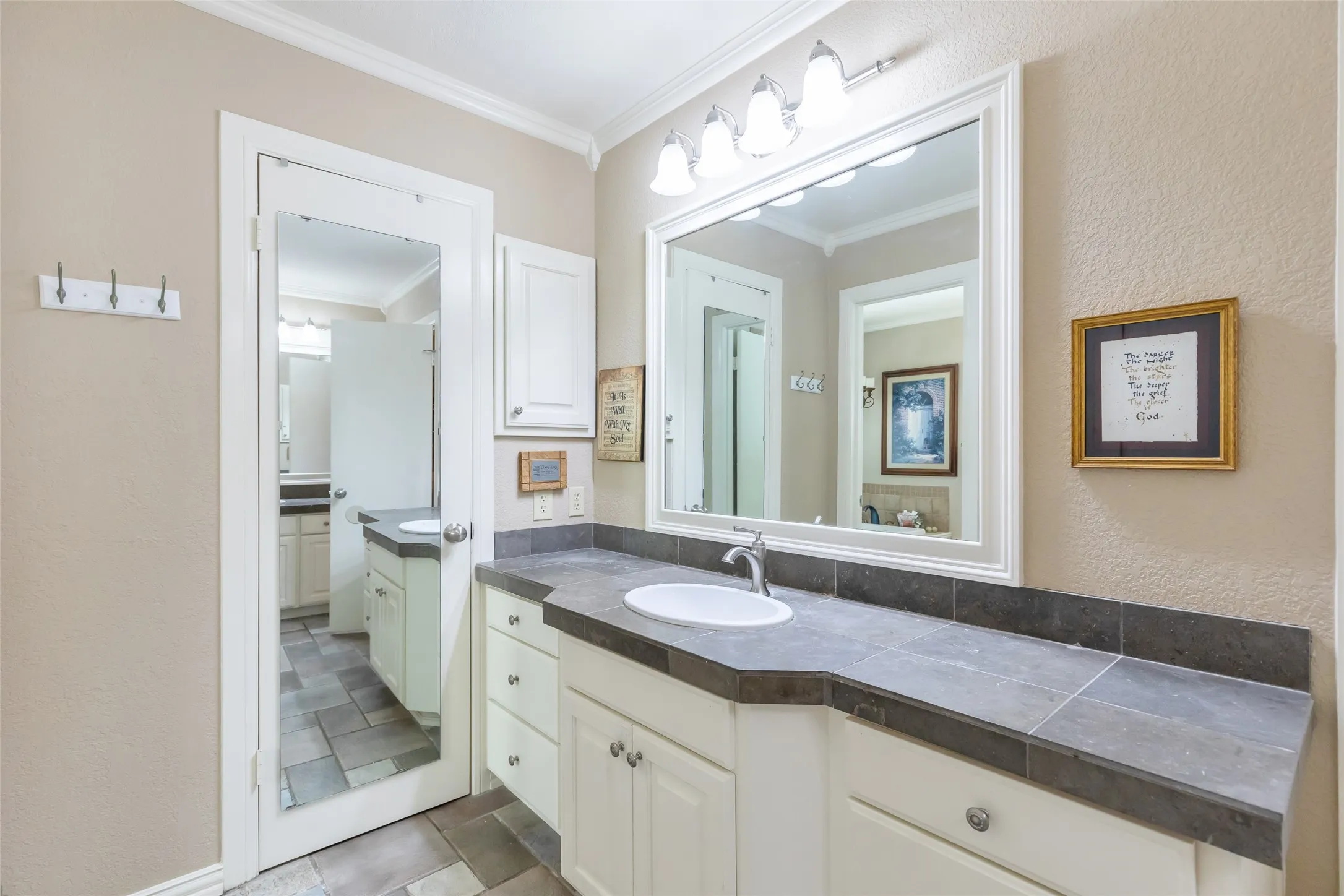 Master bathroom featuring dual vanities with cream cabinetry, dark stone countertops, and an illuminated framed mirror