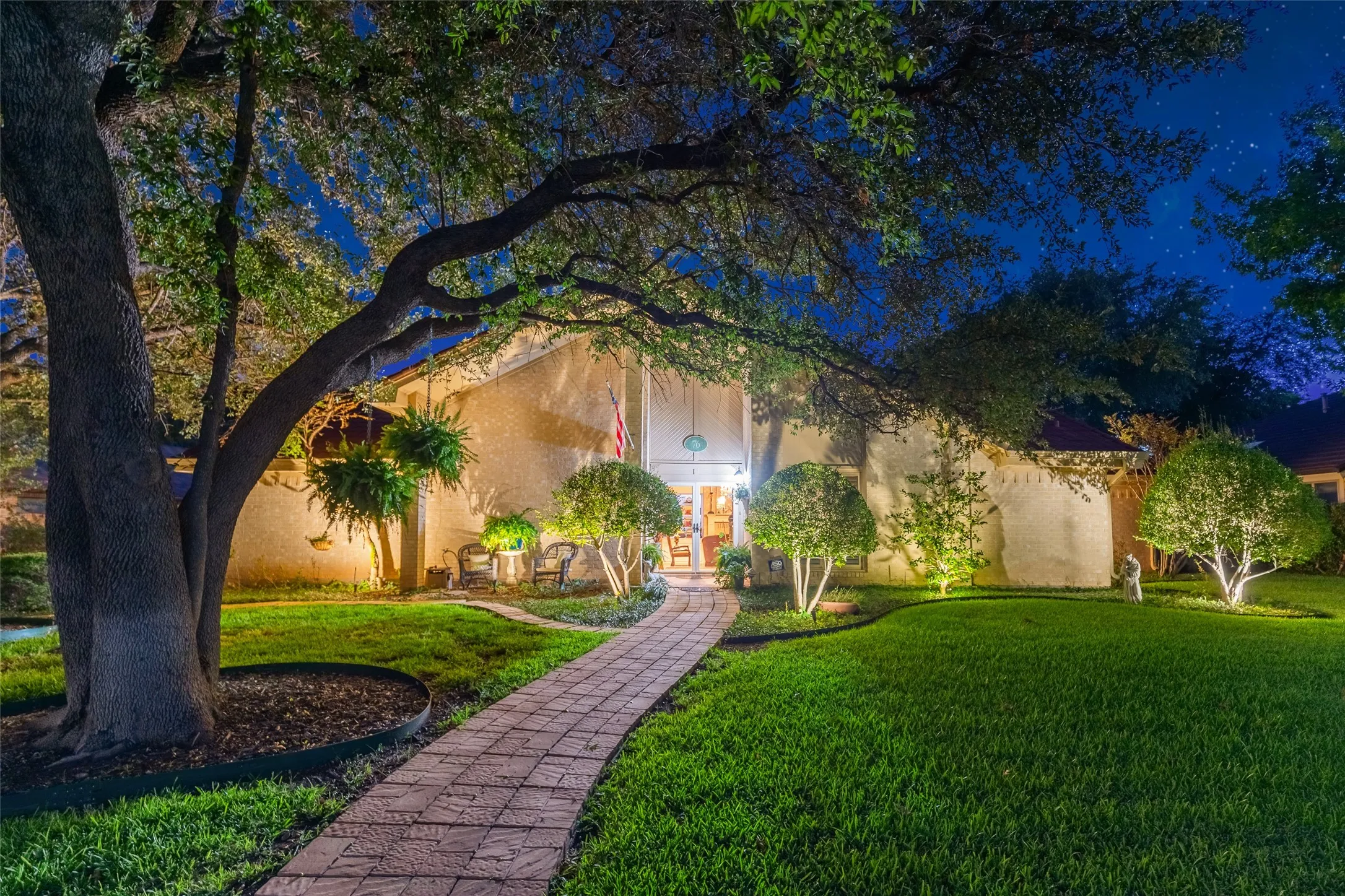 Manicured front yard featuring a paved walkway, mature trees, and landscape lighting