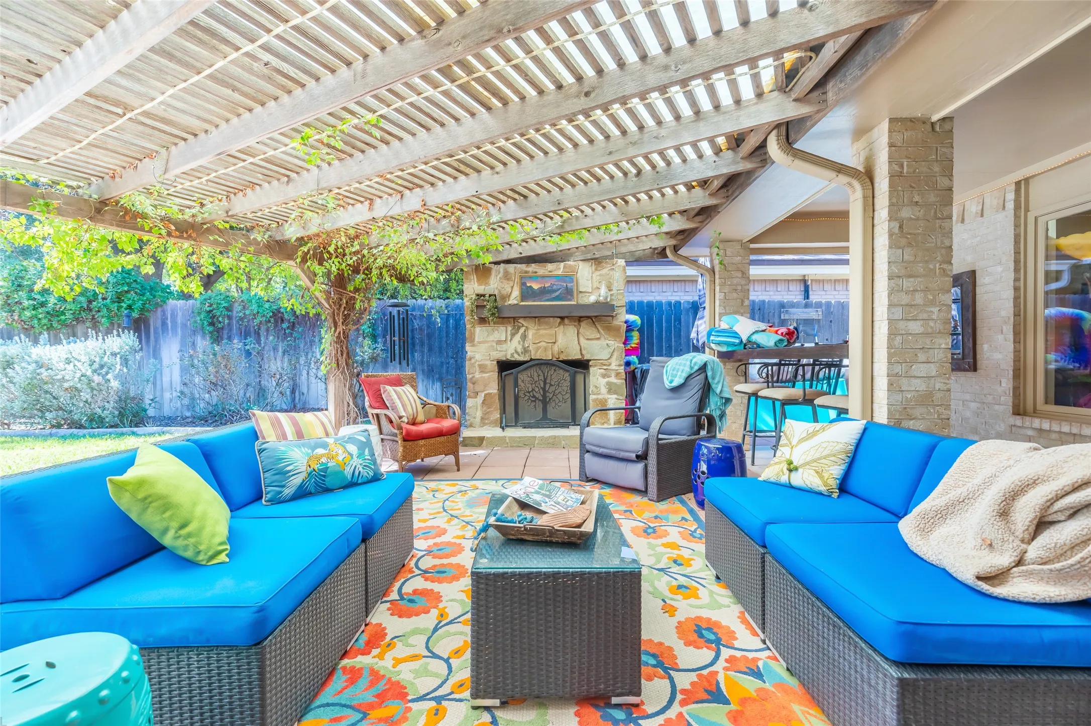 Covered patio with louvered pergola ceiling providing adjustable shade and ventilation, anchored by a dual-sided fireplace with stone surround