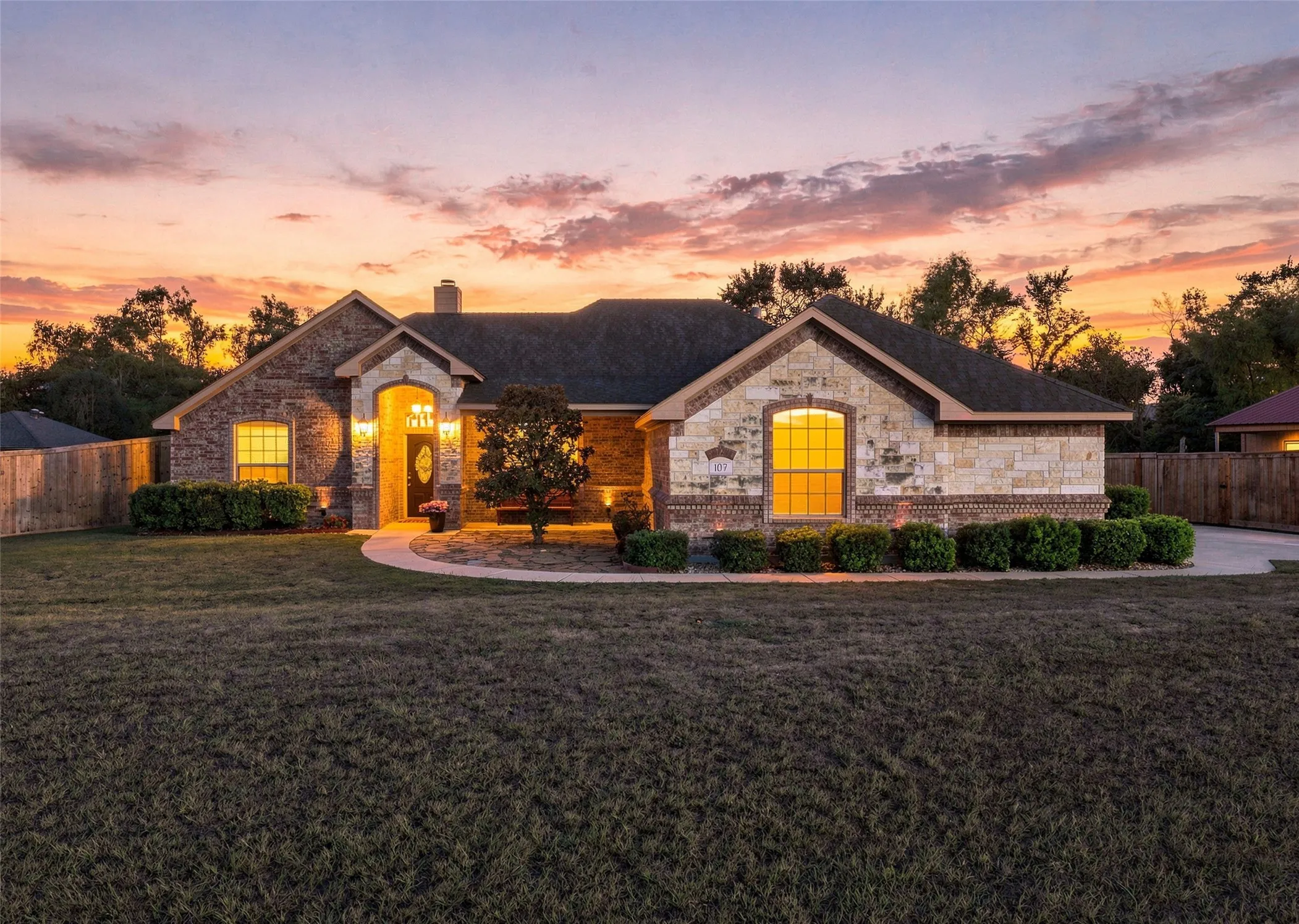 Front Exterior: Beautiful brick and stone facade highlighted by warm exterior lighting, manicured landscaping, and a colorful sunset backdrop.