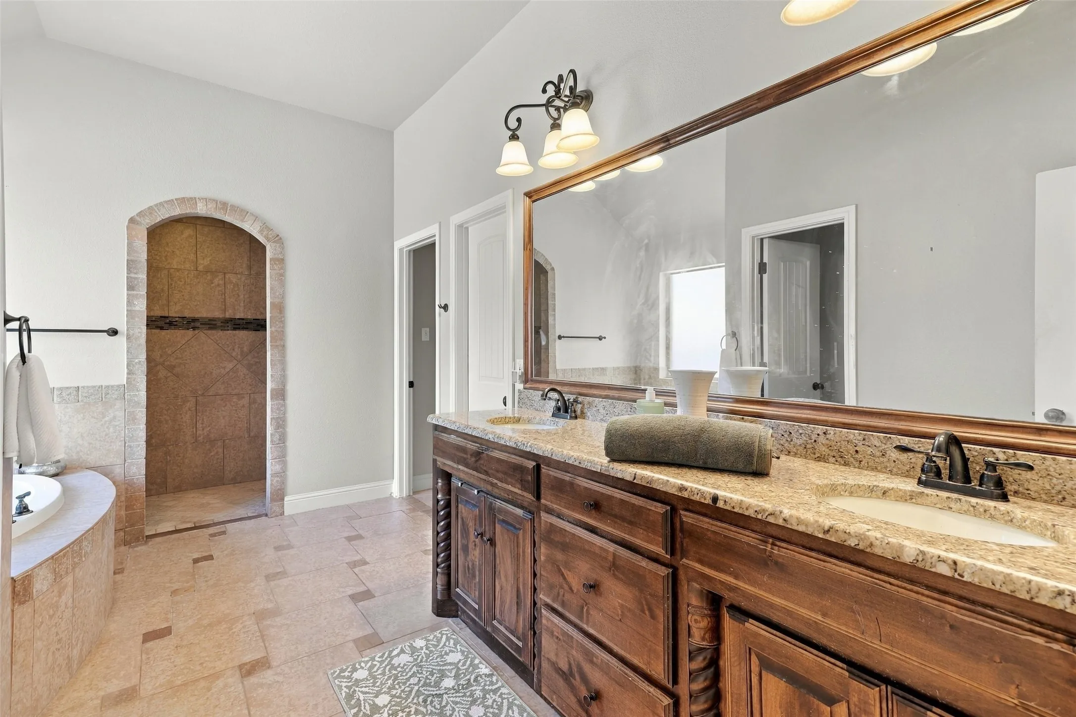 Primary Bathroom: Spacious ensuite features a granite-topped dual-sink vanity, rich wood cabinetry, decorative lighting, and a tiled walk-in shower.