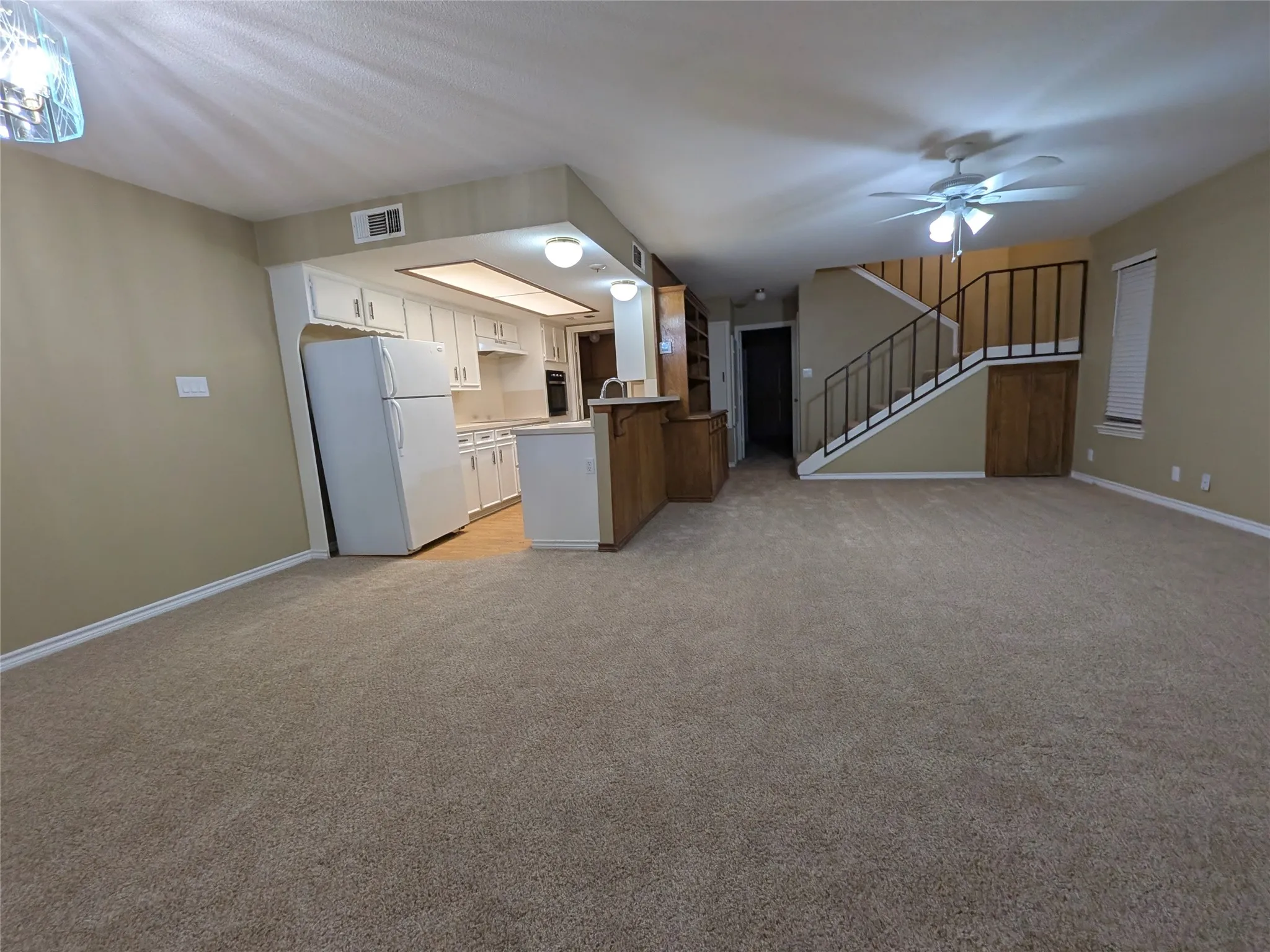Open-concept living area with carpet flooring, a kitchen featuring white cabinetry and built-in appliances, a staircase with metal railings, and a ceiling fan
