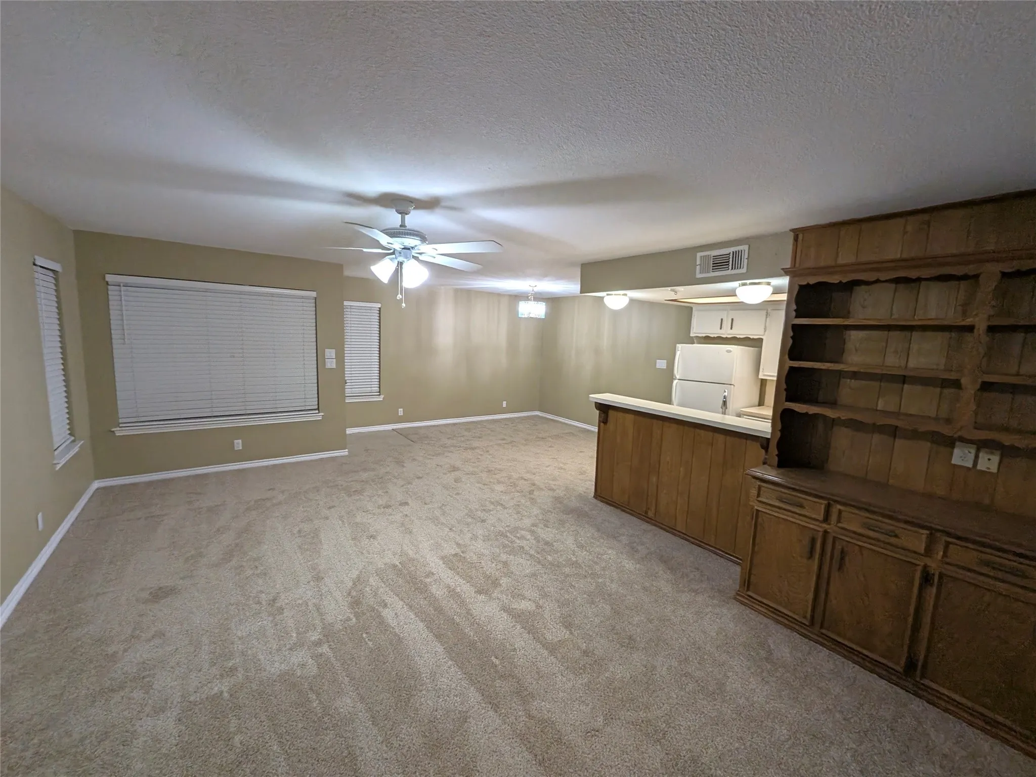 Spacious open-concept living area featuring neutral carpeting, ceiling fan with integrated lighting, built-in wood cabinetry with shelving, and a wet bar with white countertop