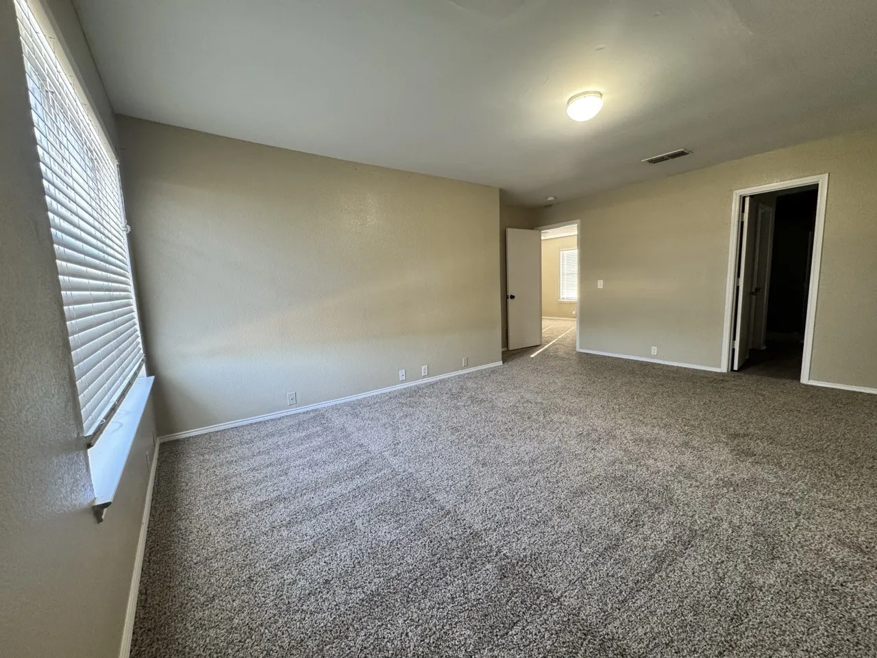 Spacious room featuring neutral wall tones, textured carpeting, a single window with blinds, and a flush mount ceiling light