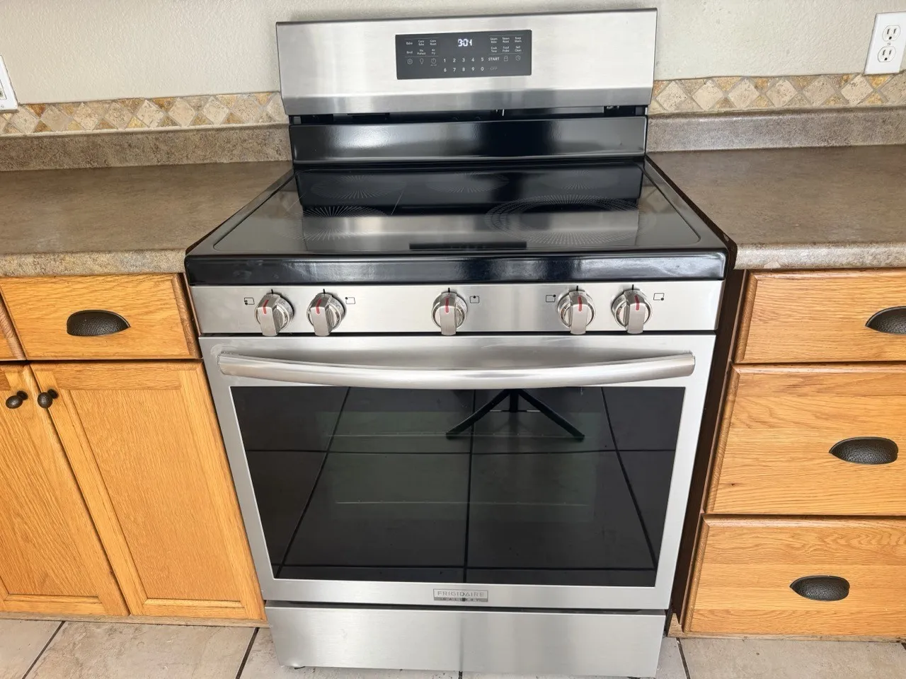 Stainless steel range, black glass cooktop, coordinating wall oven, wood-finish cabinetry, and tiled flooring
