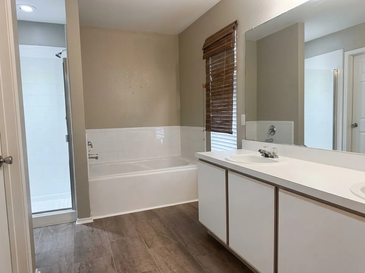 Bathroom featuring a dual vanity with integrated sinks, white cabinetry, a built-in bathtub, a walk-in shower with a glass enclosure, and wood-finish flooring