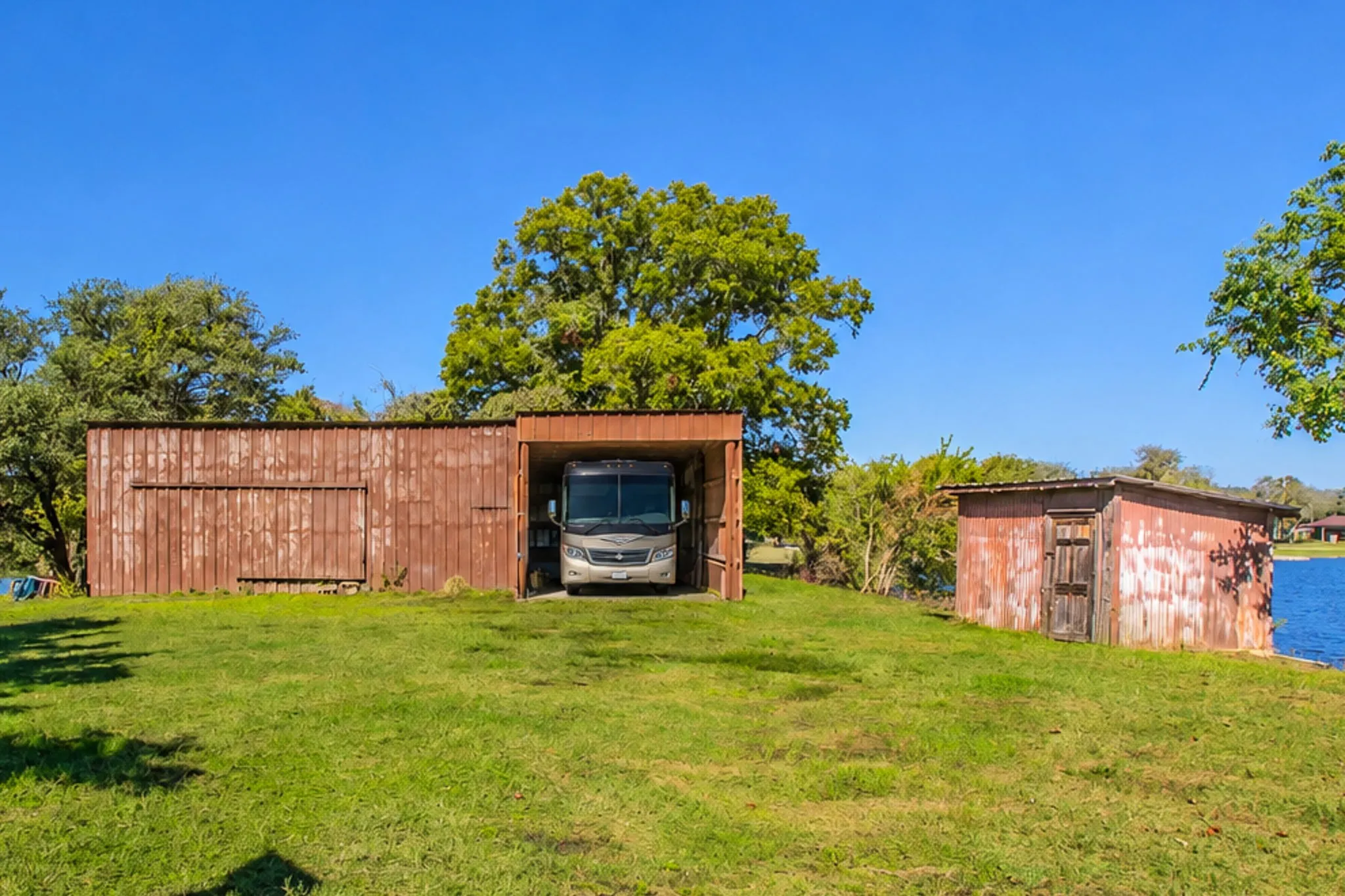 Expansive grassy property featuring a large  double level metal workshop with electricity and a RV storage area without electricity, a well shed, and mature trees. The shoreline extends behind the workshop on both sides.