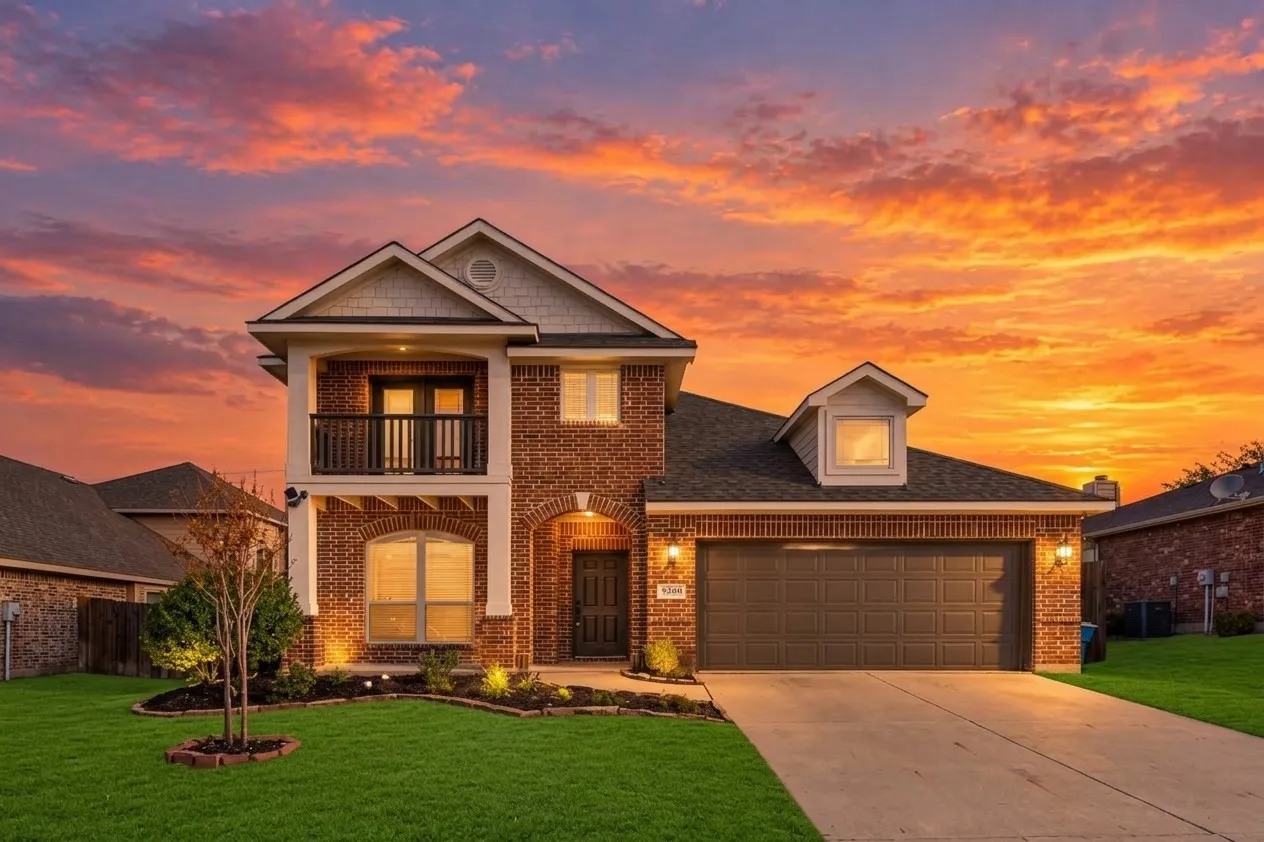 Brick exterior with a second-story balcony featuring dark railing, an arched entry, and an attached two-car garage with a panel door. Grass edited greener with AI