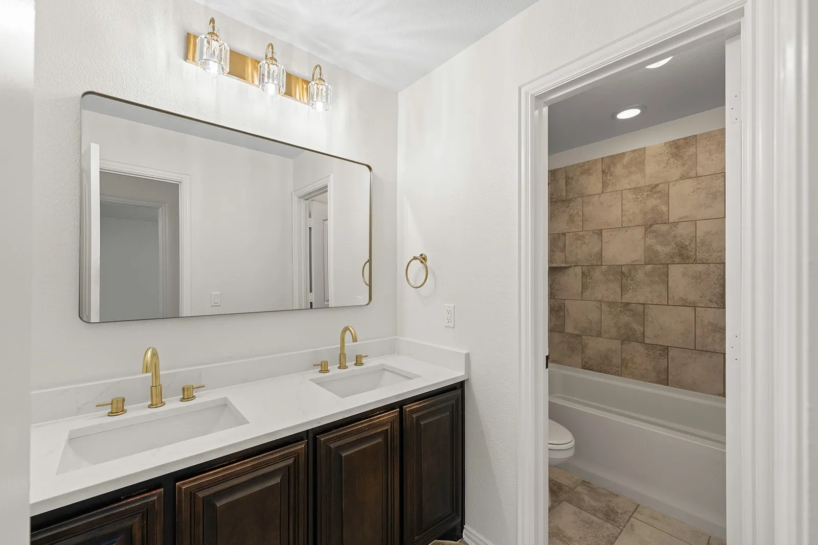 Dual vanity with gold-finish fixtures and hardware, rectangular mirror, and an adjacent bath with tiled walls