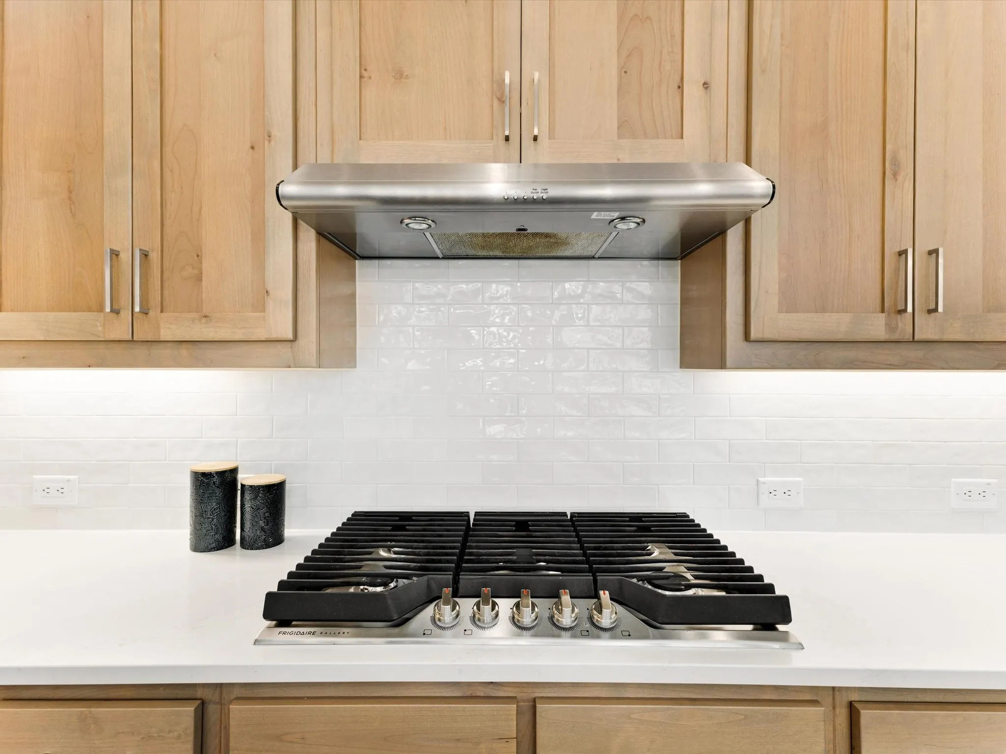 Stainless steel range hood, white subway tile backsplash, light wood cabinetry, built-in cooktop, and light-toned countertop