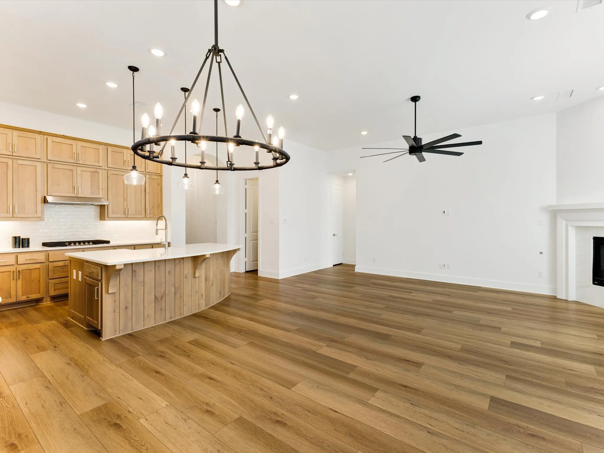 Open-concept living space with wood-finish flooring, a kitchen island with seating, natural wood cabinetry, and a fireplace with a white mantel