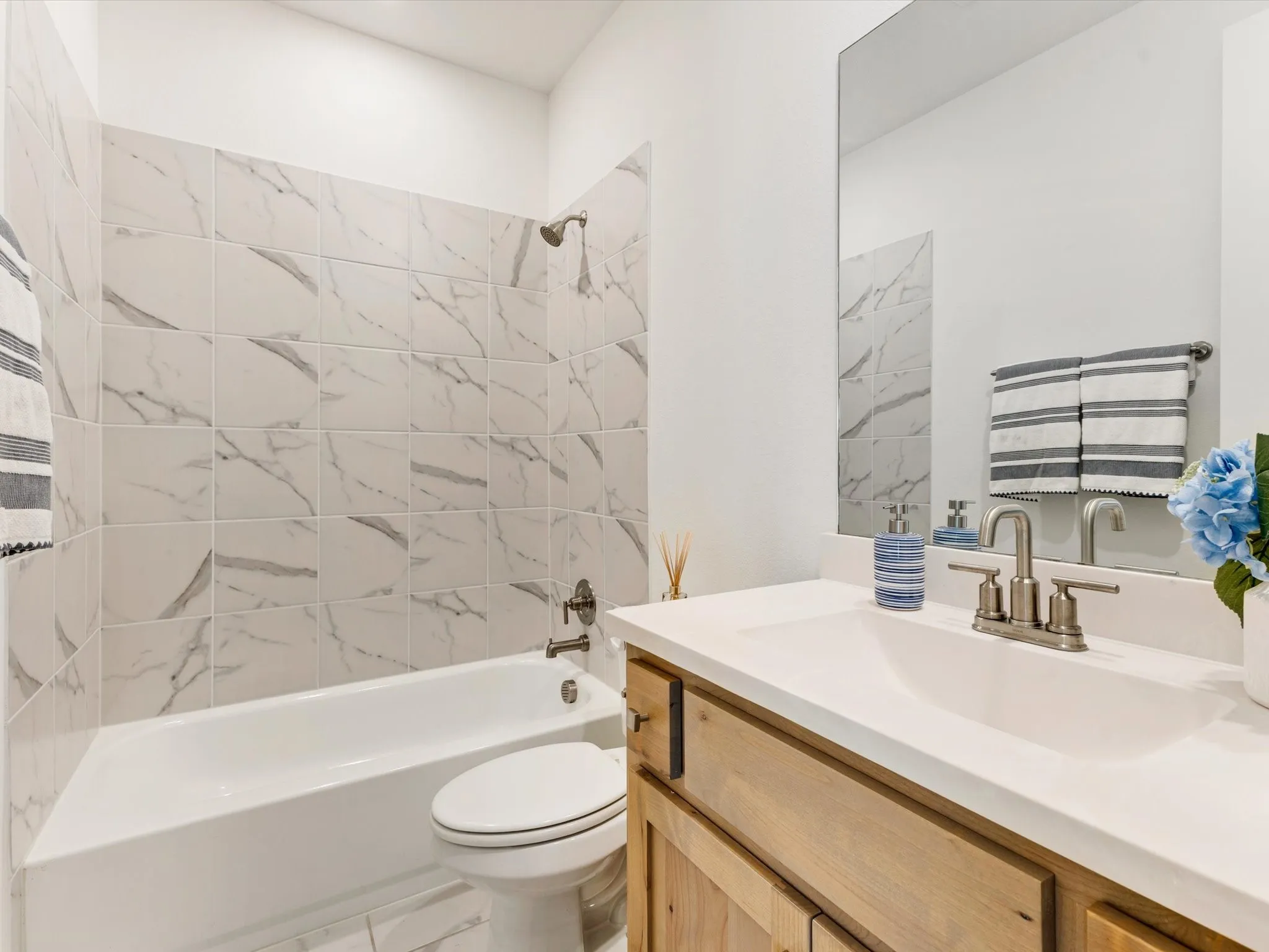 White bathtub with a shower, marble-patterned wall tile, and a wood-finish vanity with a white countertop and undermount sink