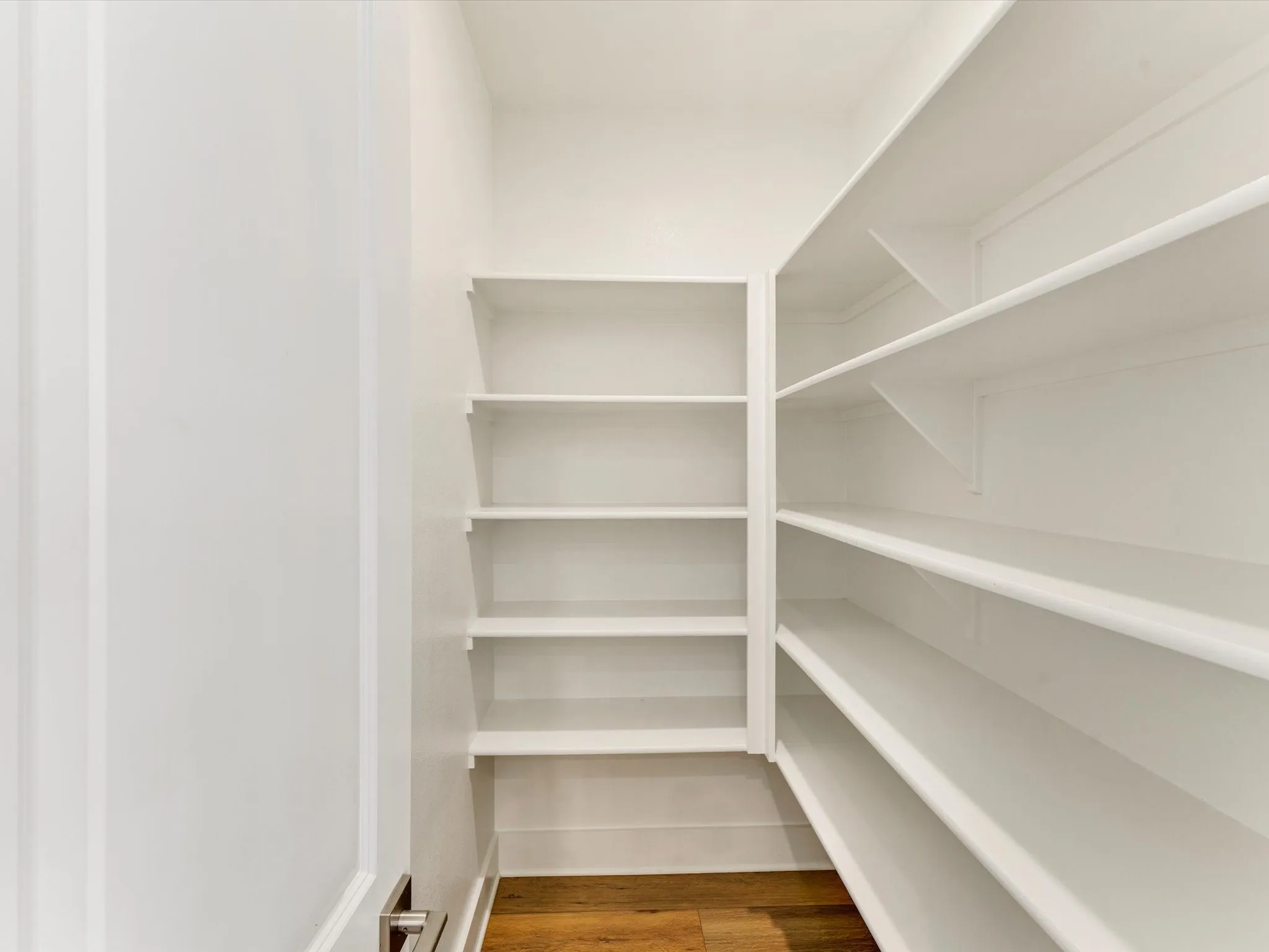 Ample pantry storage with multiple built-in white shelving units, wood-finish flooring, and crisp white walls