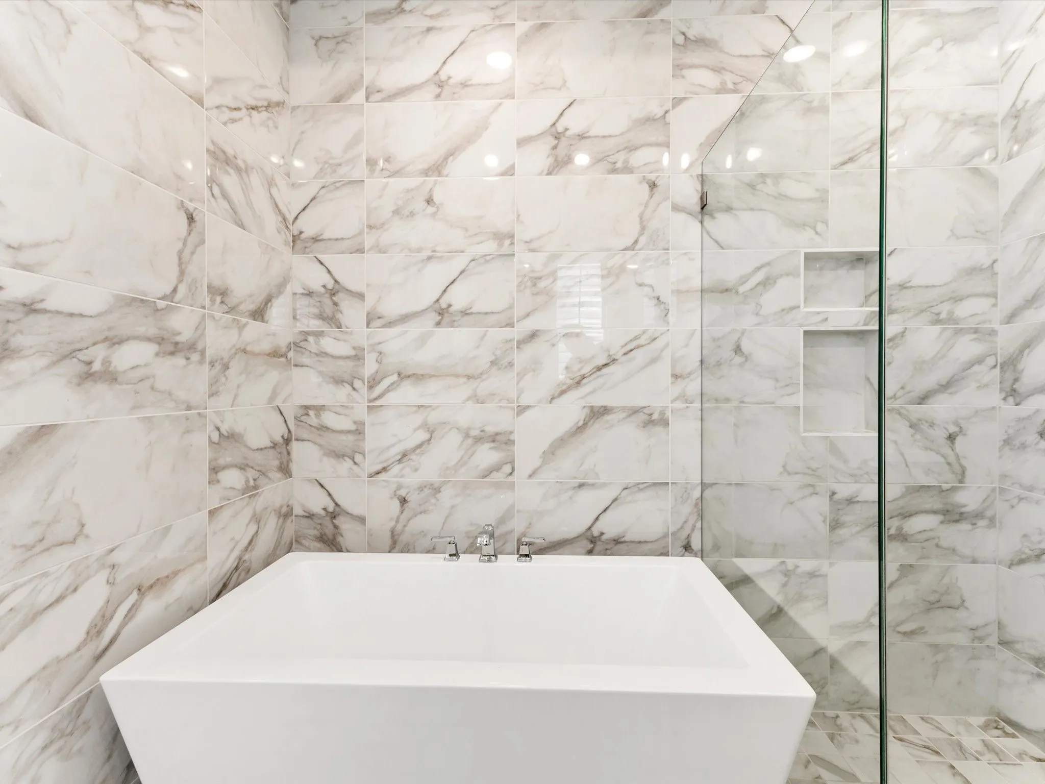 Freestanding soaking tub with polished chrome fixtures, surrounded by large format tile with natural stone veining, and a frameless glass shower enclosure featuring built-in niches