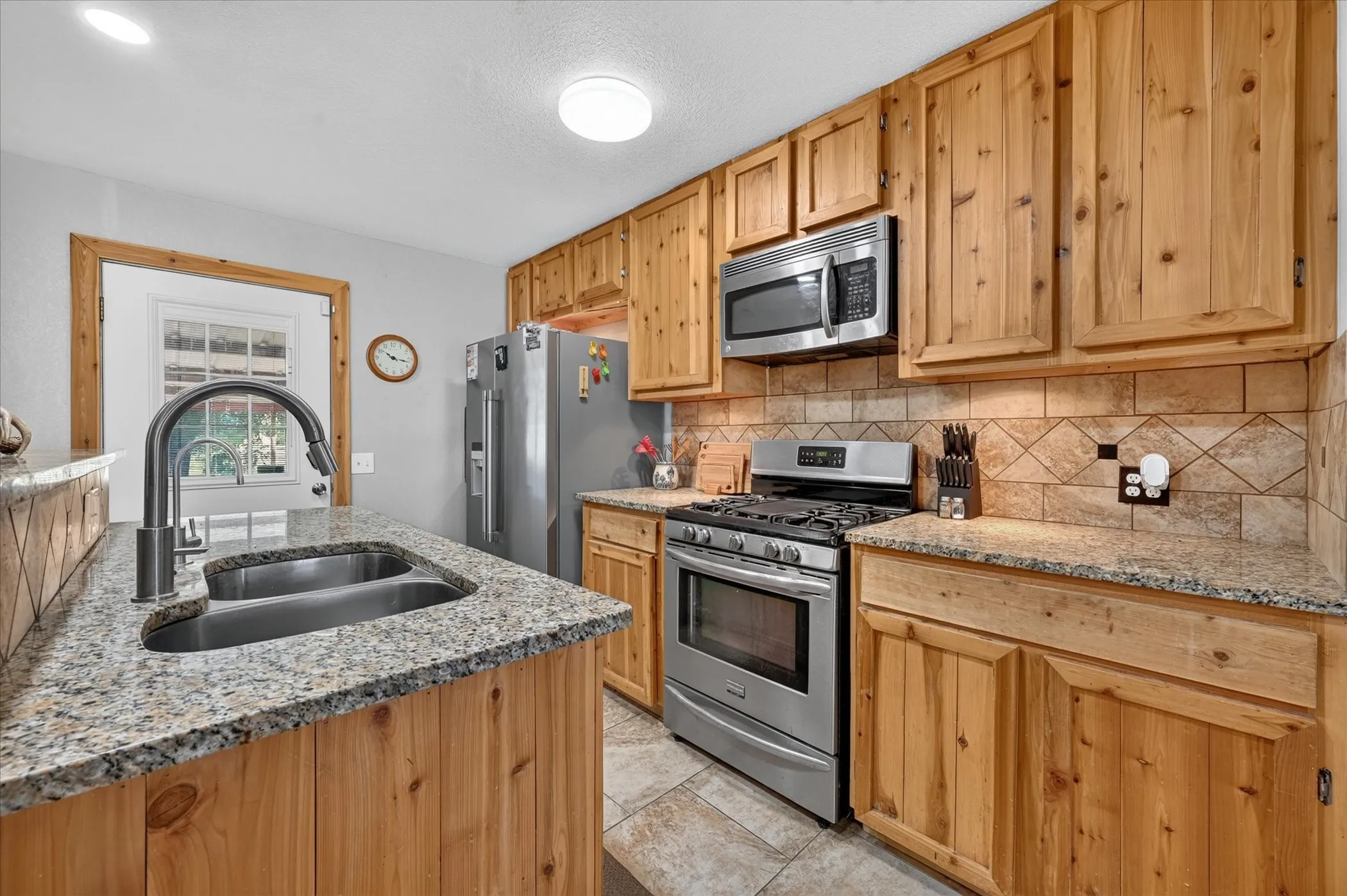 Kitchen featuring wood cabinetry, stainless steel appliances, granite-style countertops, tile backsplash, and a double basin sink with a gooseneck faucet