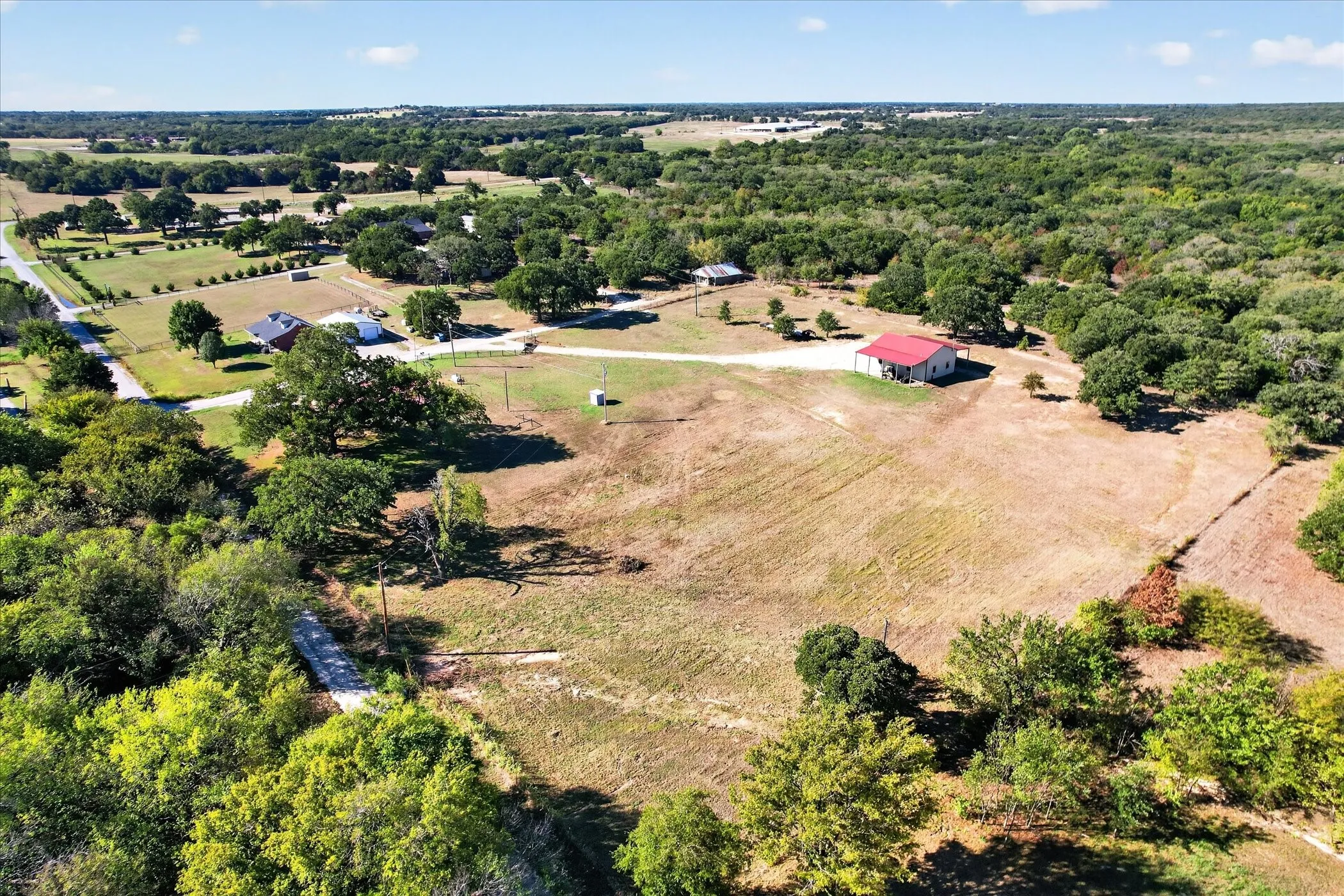 Sprawling acreage featuring a mature tree line, a detached building with a red roof, and a gravel driveway