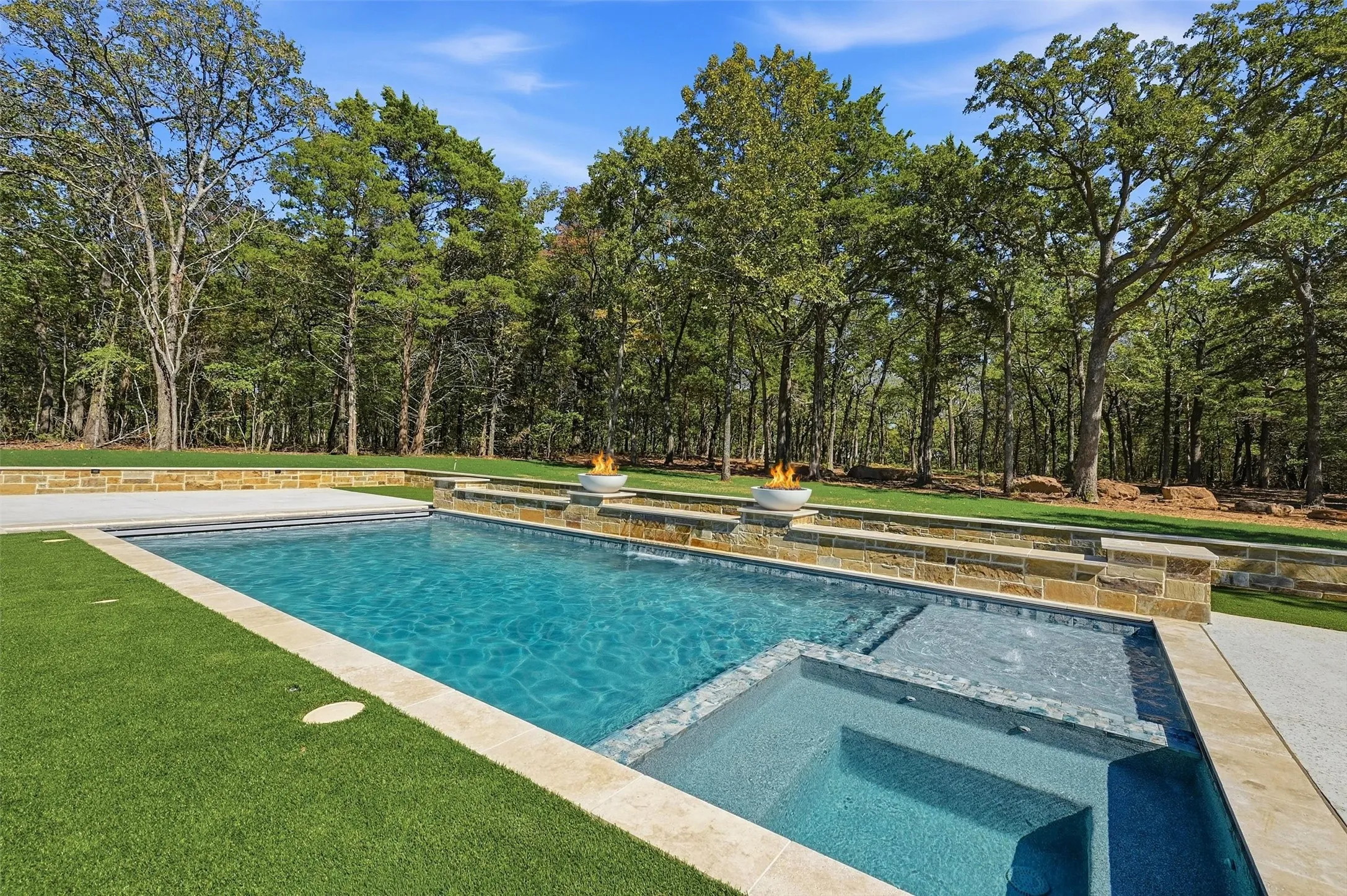 In-ground swimming pool with an integrated spa, stone-clad retaining wall, artificial turf, and mature tree-lined perimeter