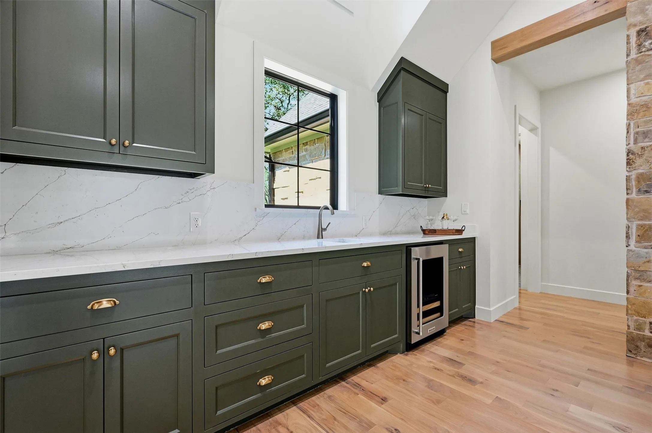 Built-in cabinetry with brass hardware, integrated beverage cooler, stone countertops with matching backsplash, wood-finish flooring, and a visible wood beam