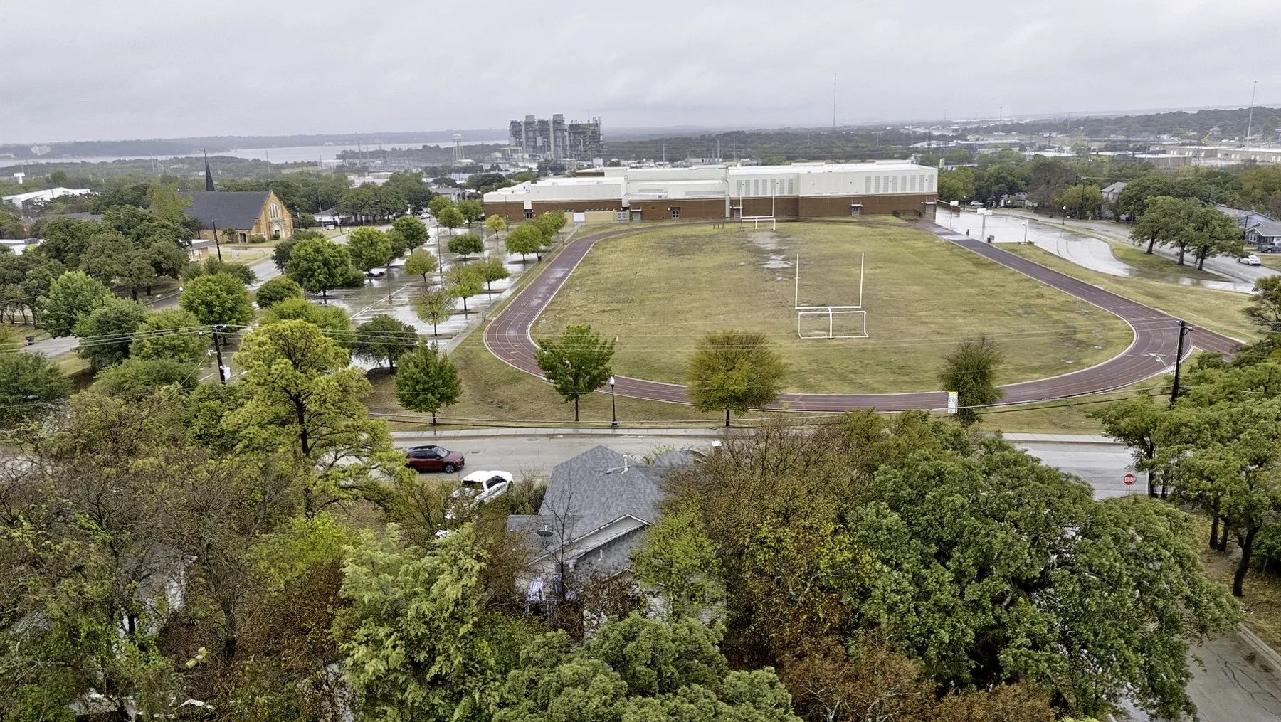 Aerial of the home's gray shingle roof with the track and field across the street and a lake on the far horizon