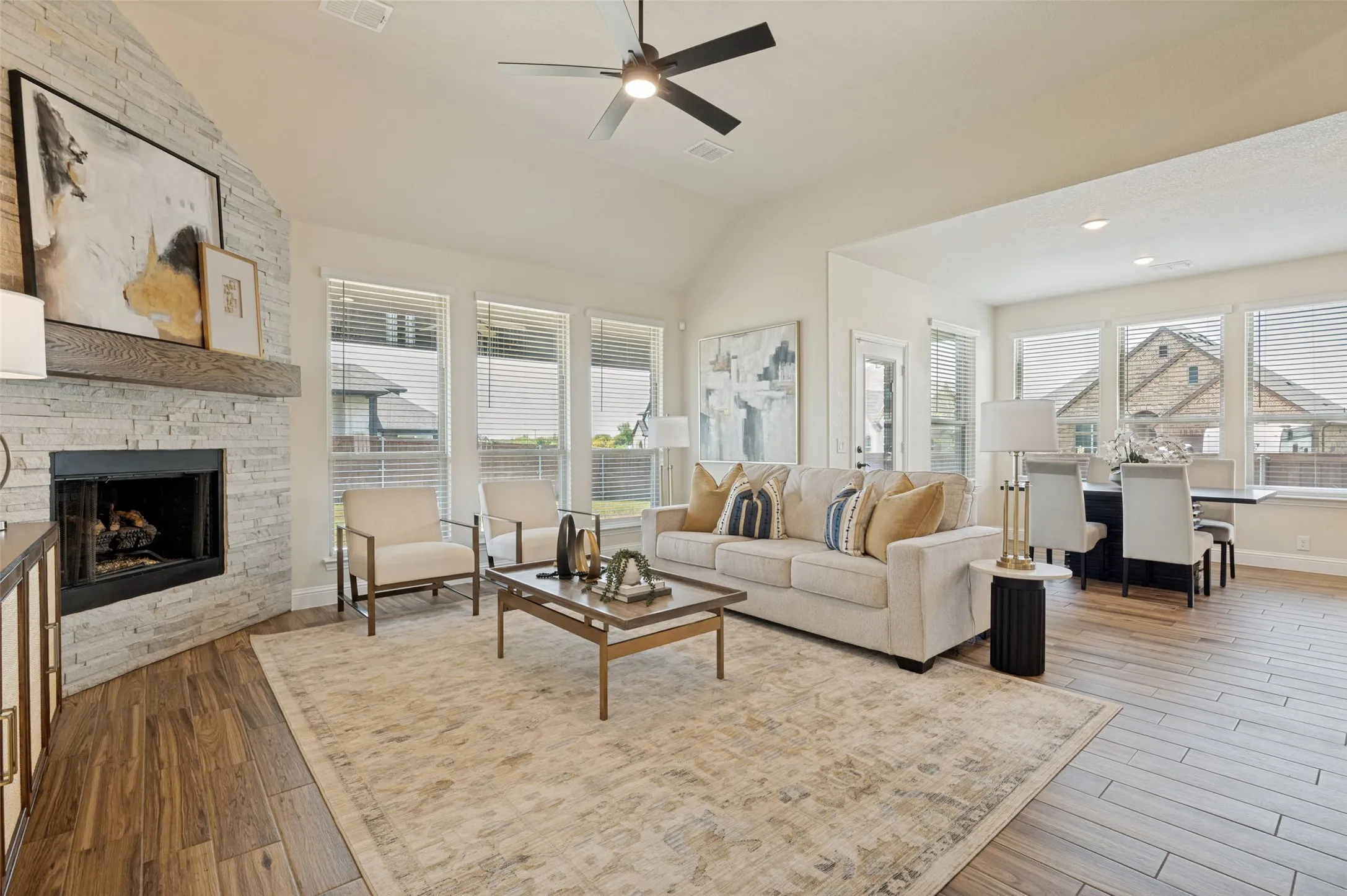 Vaulted ceilings, wood-finish flooring, stone-clad fireplace with wood mantle, multiple windows with blinds, and recessed lighting