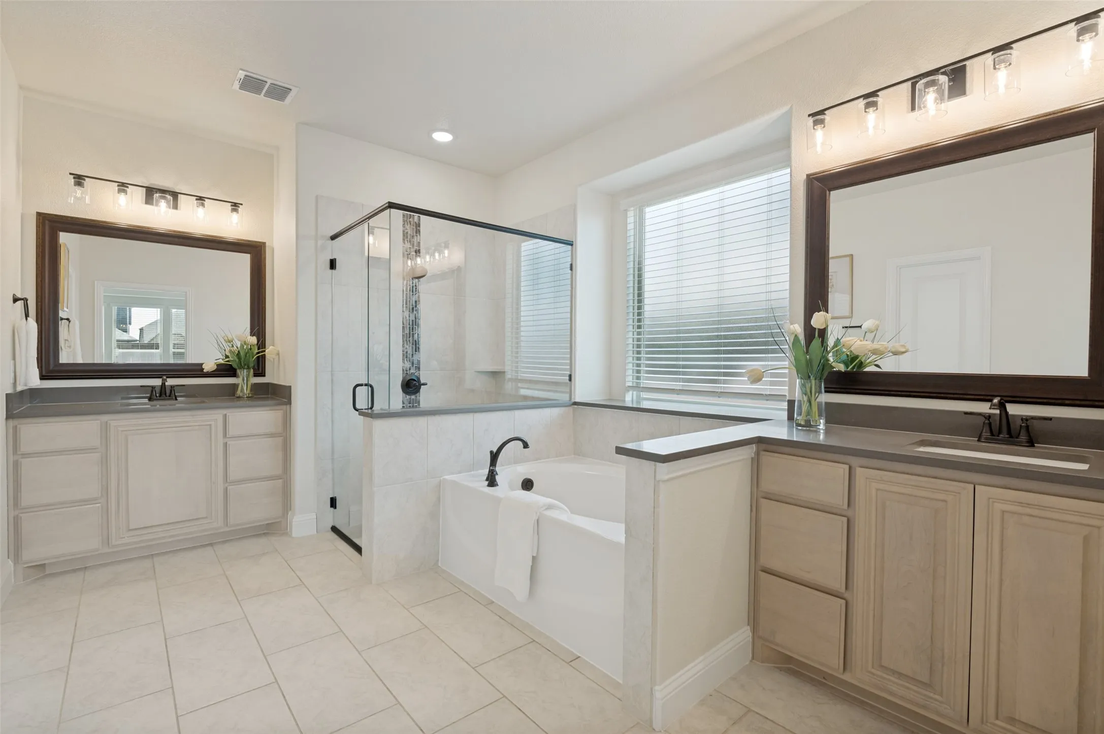 Dual vanity sinks with dark countertops, enclosed glass shower, built-in bathtub, and a large window with blinds