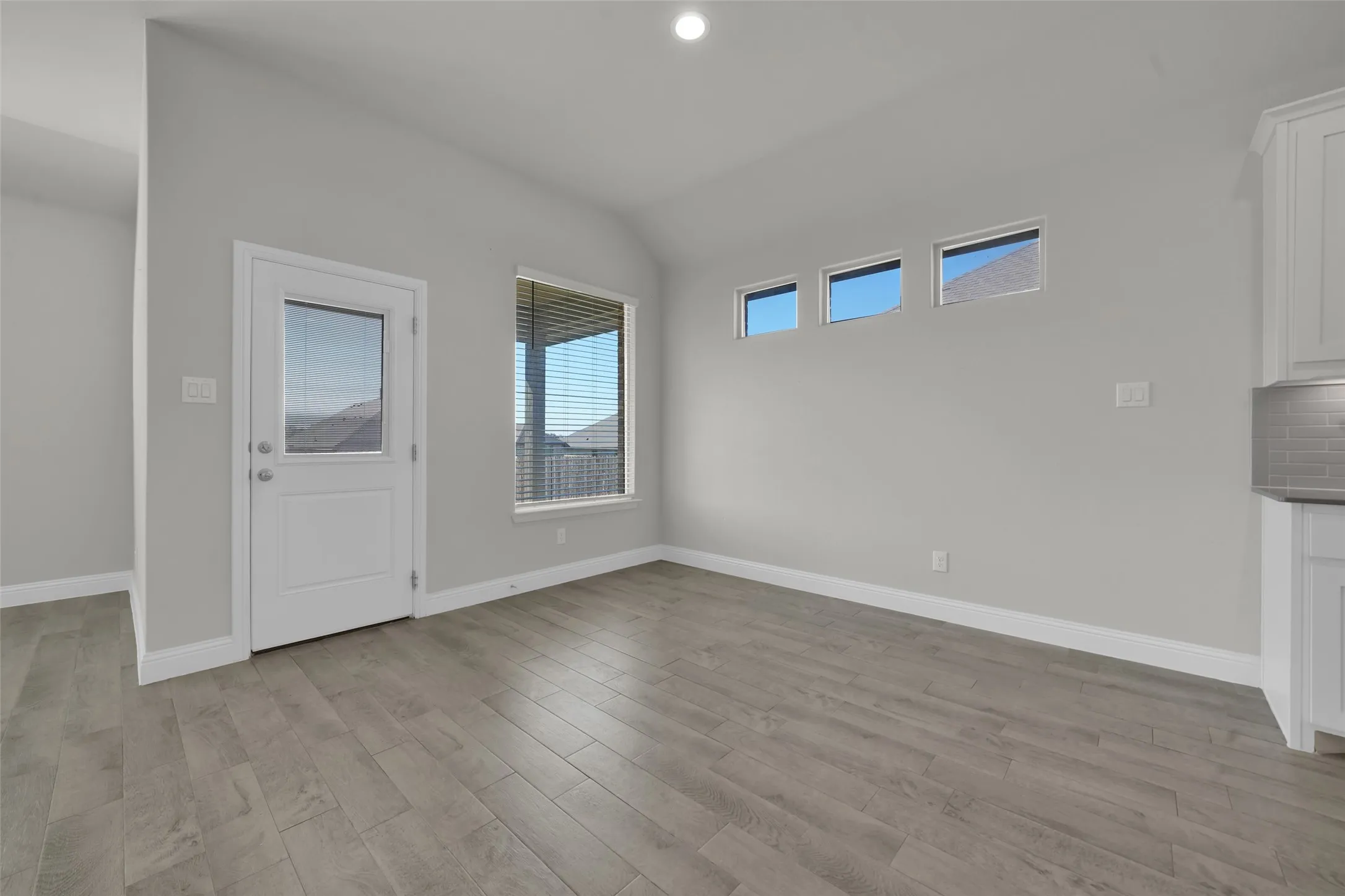 Vaulted ceiling, wood-finish flooring, glass-paned exterior door, multiple windows, and partial kitchen cabinetry with subway tile backsplash