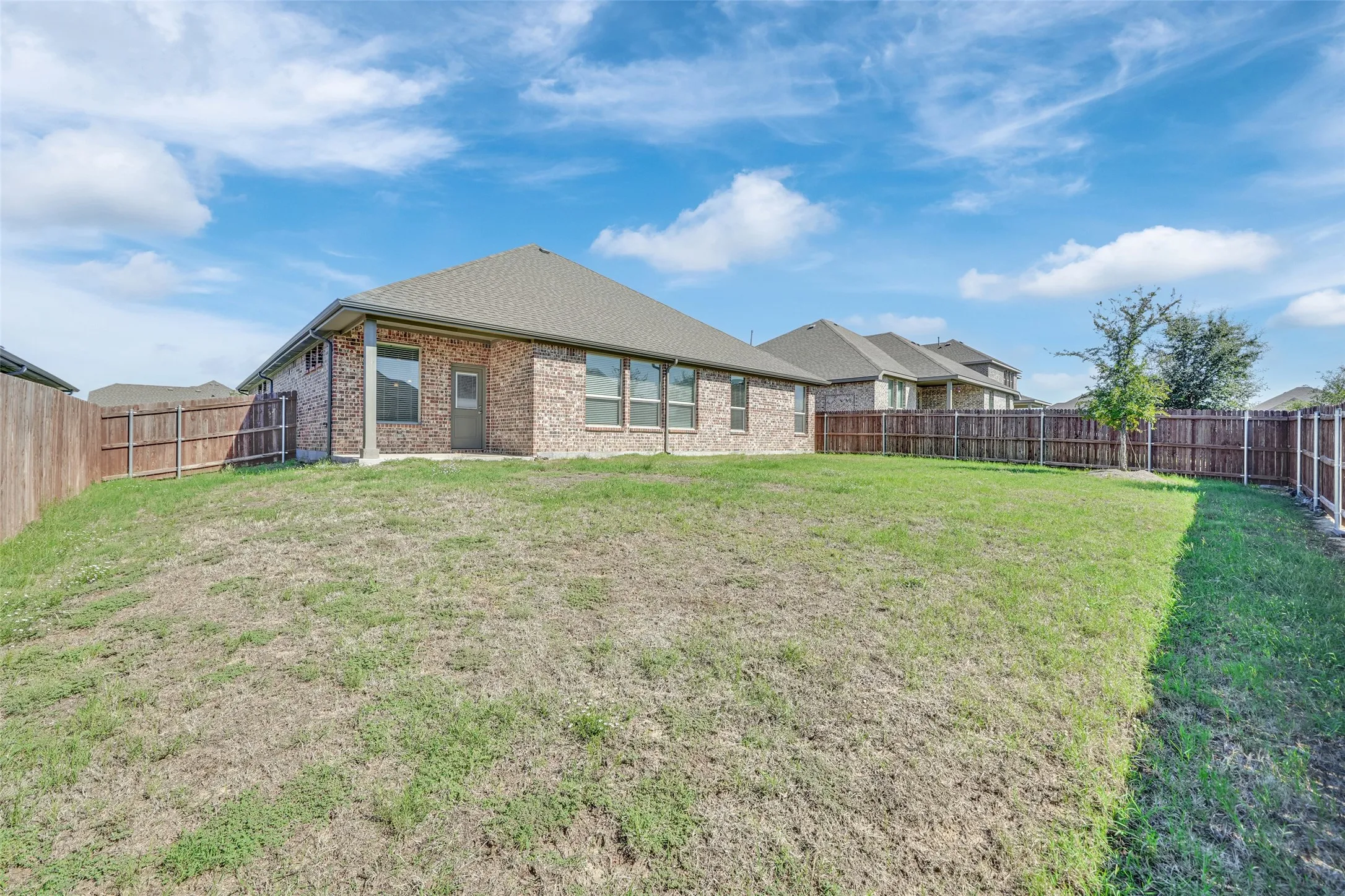 Expansive backyard featuring a privacy fence, brick exterior, multiple windows, and a paved patio area