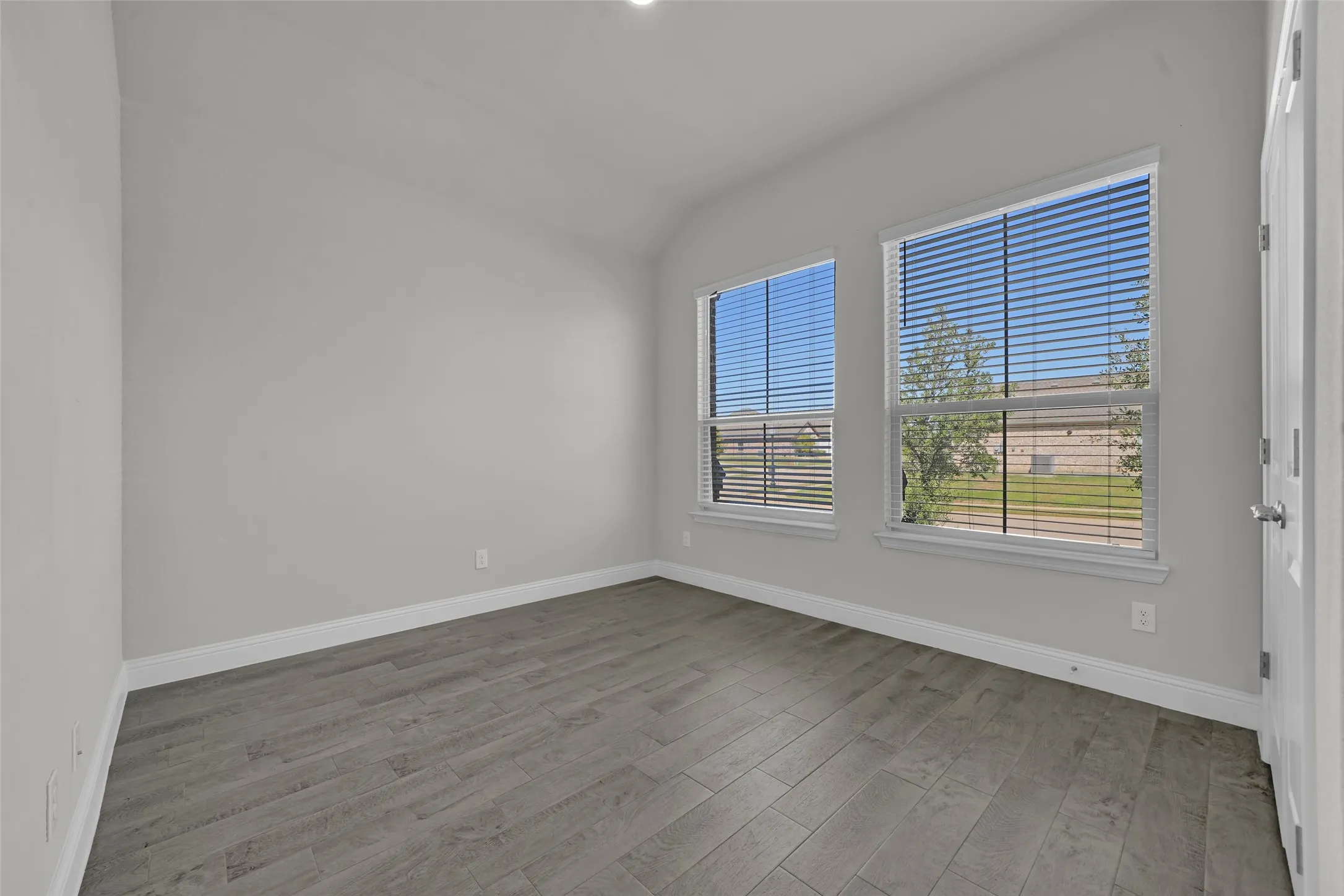 Light gray wall paint, wood-finish flooring, two windows with blinds, and a white door with a silver handle
