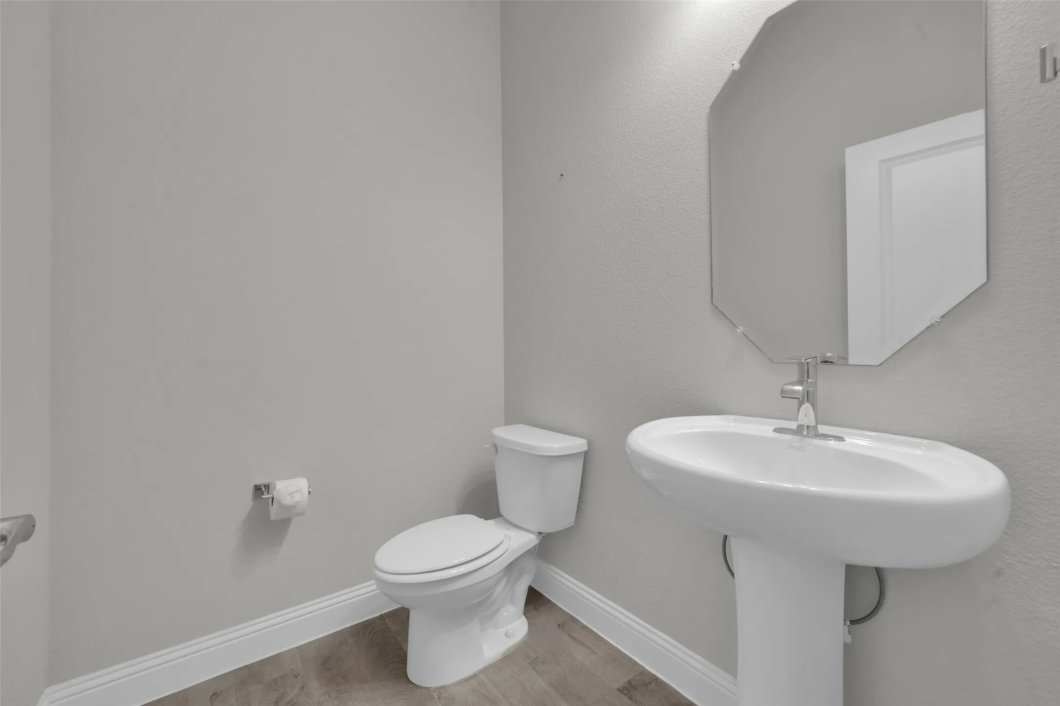 Pedestal sink with a polished chrome faucet, octagonal frameless mirror, and wood-finish flooring