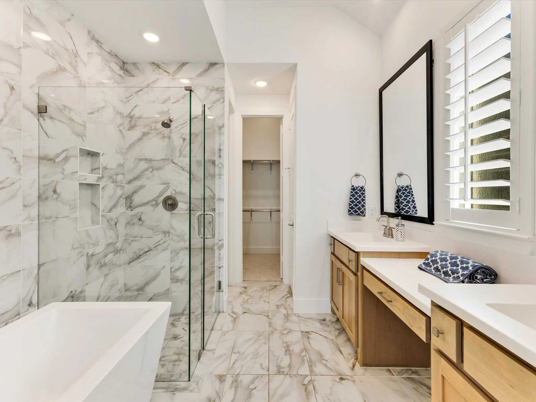 Freestanding soaking tub, walk-in shower with glass enclosure and recessed shelving, dual vanity with wood cabinetry, and large format marble-look tile flooring