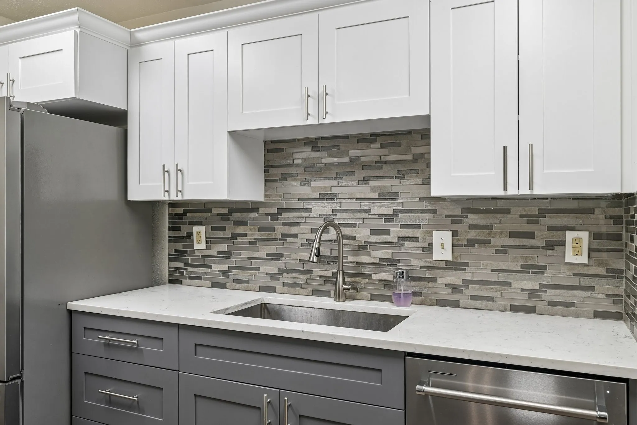 Two-tone cabinetry with white upper and gray lower cabinets, light-toned countertops, a stainless steel sink with a gooseneck faucet, and a horizontal mosaic tile backsplash