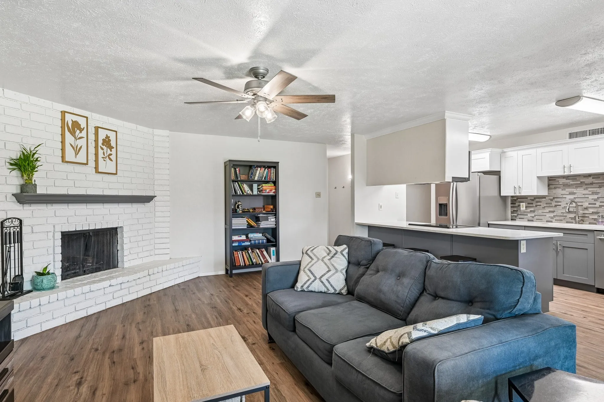 Wood-finish flooring, white brick fireplace with dark mantel, ceiling fan with integrated lighting, built-in open shelving, and a kitchen featuring white cabinetry and stainless steel appliances
