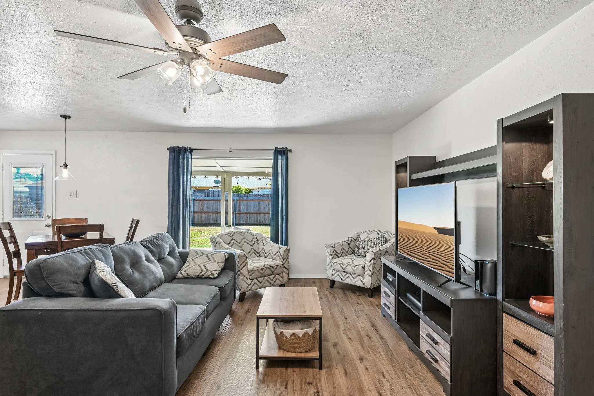 Wood-finish flooring throughout, ceiling fan with integrated lighting, built-in entertainment unit with shelving, sliding glass door leading to the exterior, and a dining area with a pendant light fixture