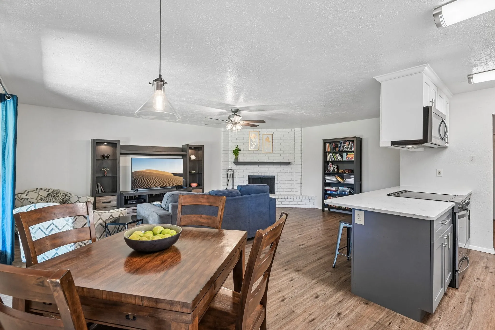 Open-concept living space featuring wood-finish flooring, a white brick fireplace, and a kitchen island with built-in cooktop