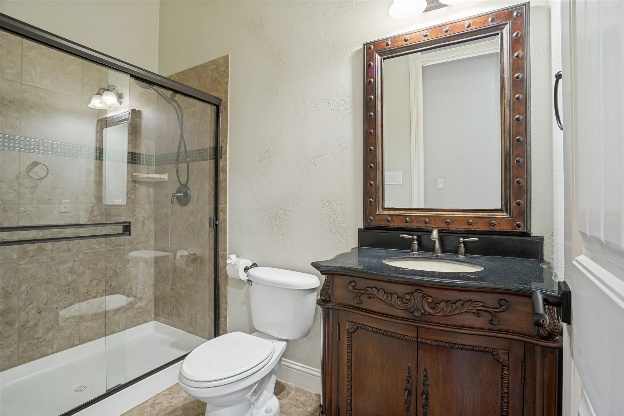 Frameless glass shower enclosure, decorative shower tile inlay, vanity with carved wood details, dark countertop, and decorative framed mirror