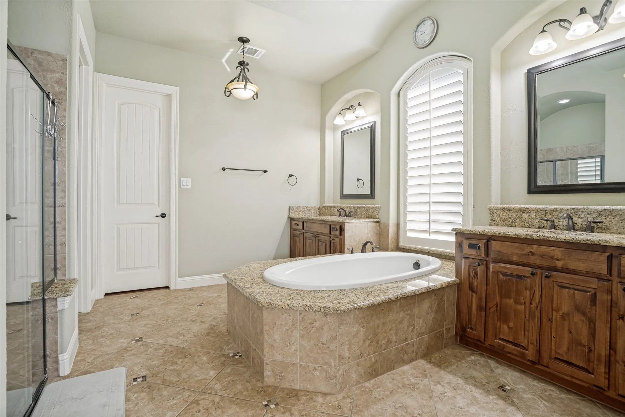 Spacious bathroom featuring a built-in soaking tub with a stone surround, dual wood vanities with stone countertops, and an arched window with plantation shutters