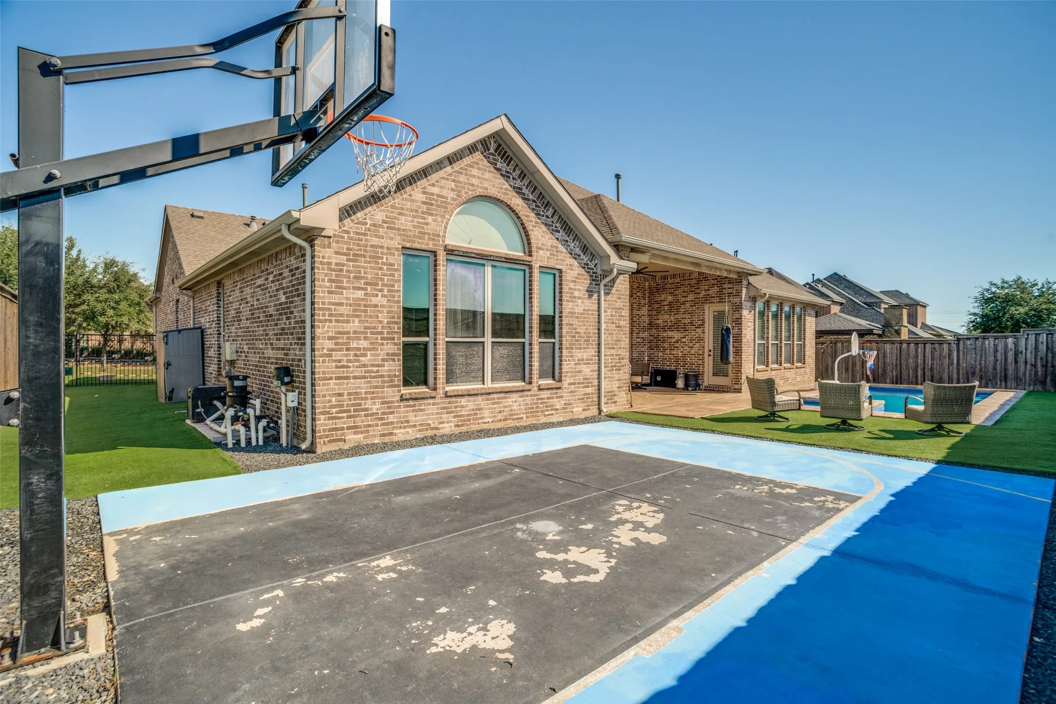 Brick exterior with arched windows, a covered patio, and a fenced backyard featuring a paved sports court, artificial turf, and an in-ground pool