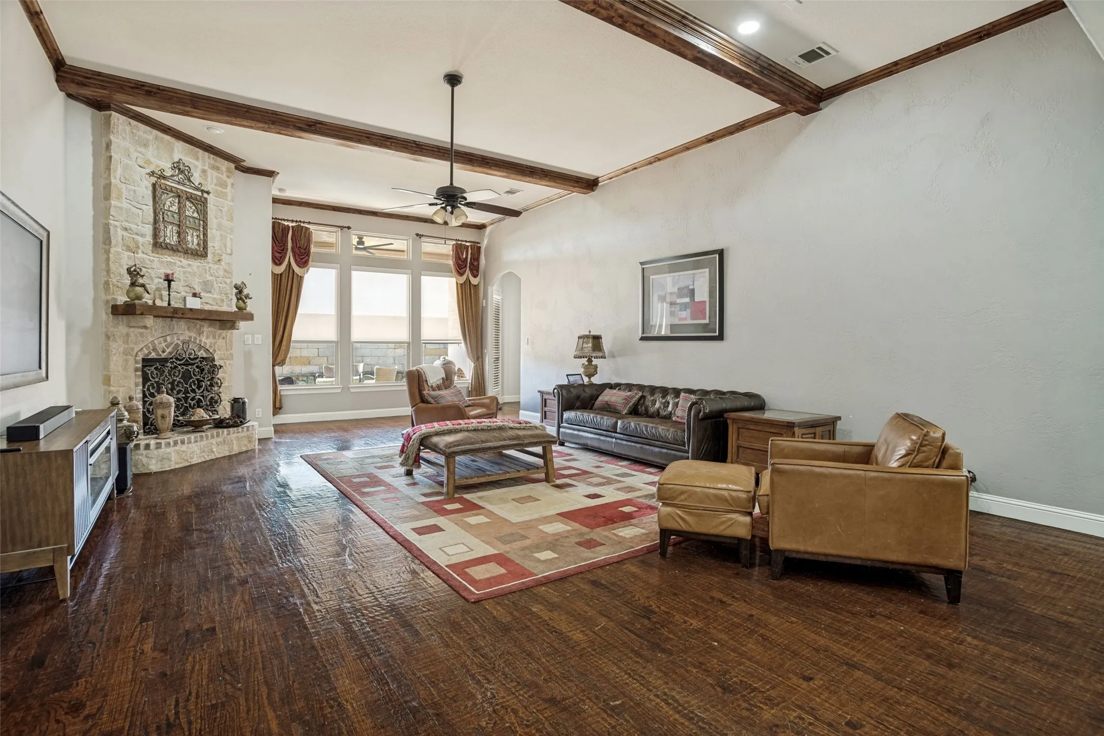 Vaulted ceiling with exposed wood beams, stone-clad fireplace with mantel, dark wood-finish flooring, and large windows