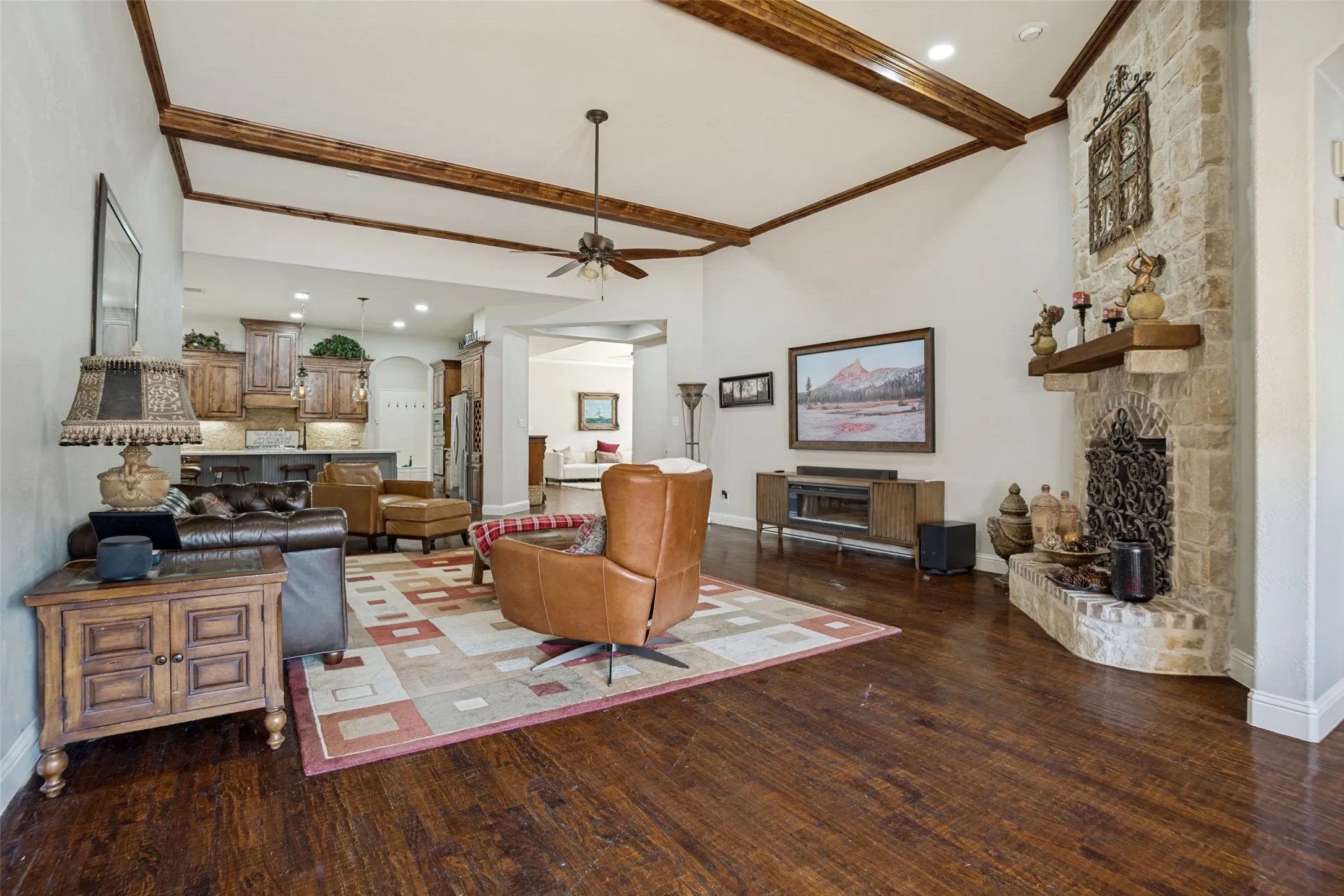 Vaulted ceiling with exposed wood beams, stone-clad fireplace with hearth, dark wood-finish flooring, and recessed lighting