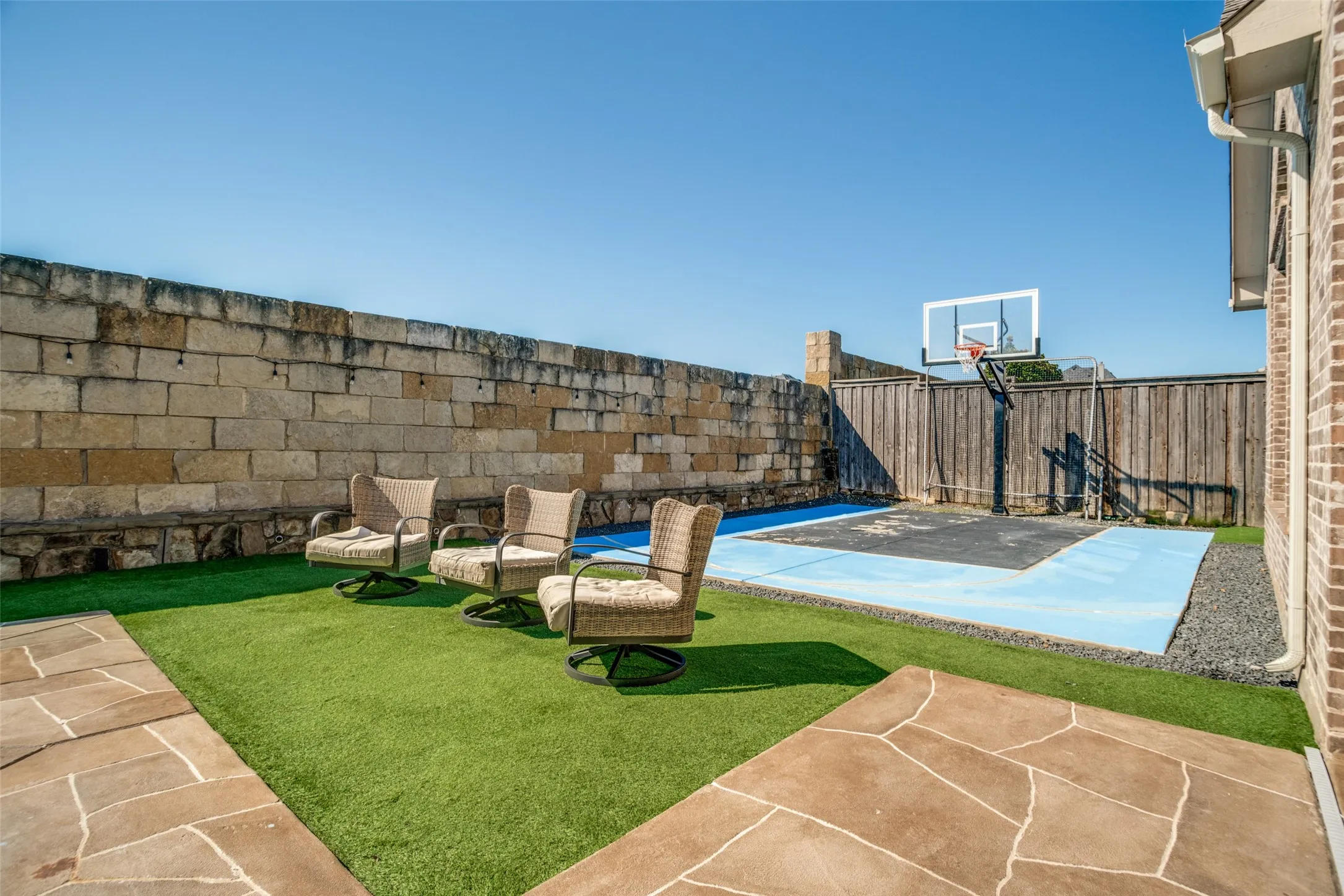 Backyard featuring a stone retaining wall, artificial turf, flagstone-style patio, and a dedicated court with a basketball hoop