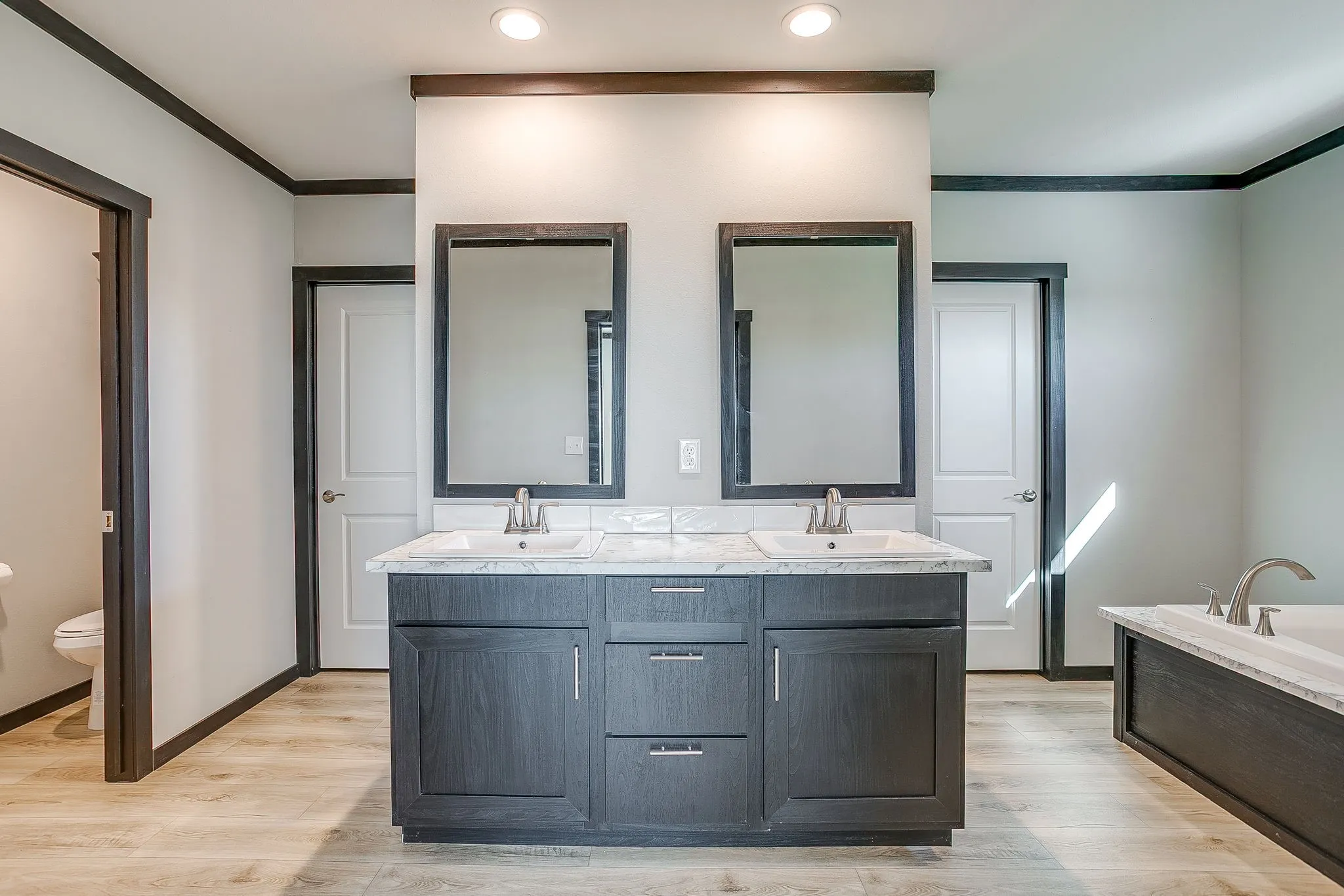 Dual vanity with integrated sinks, wood-finish cabinetry, light-toned countertops, wood-finish flooring, and a built-in bathtub