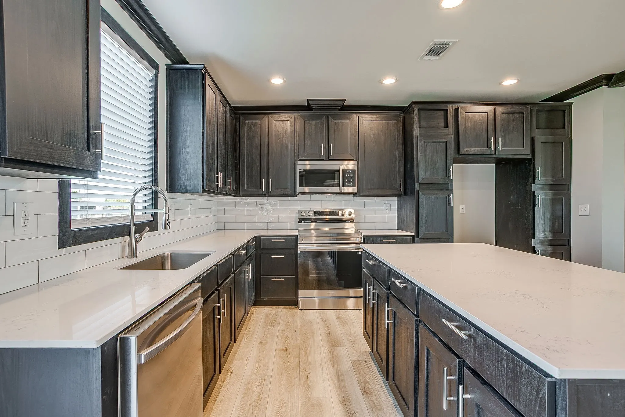 Kitchen featuring dark cabinetry, stainless steel appliances, a large island with a light-toned countertop, subway tile backsplash, and wood-finish flooring