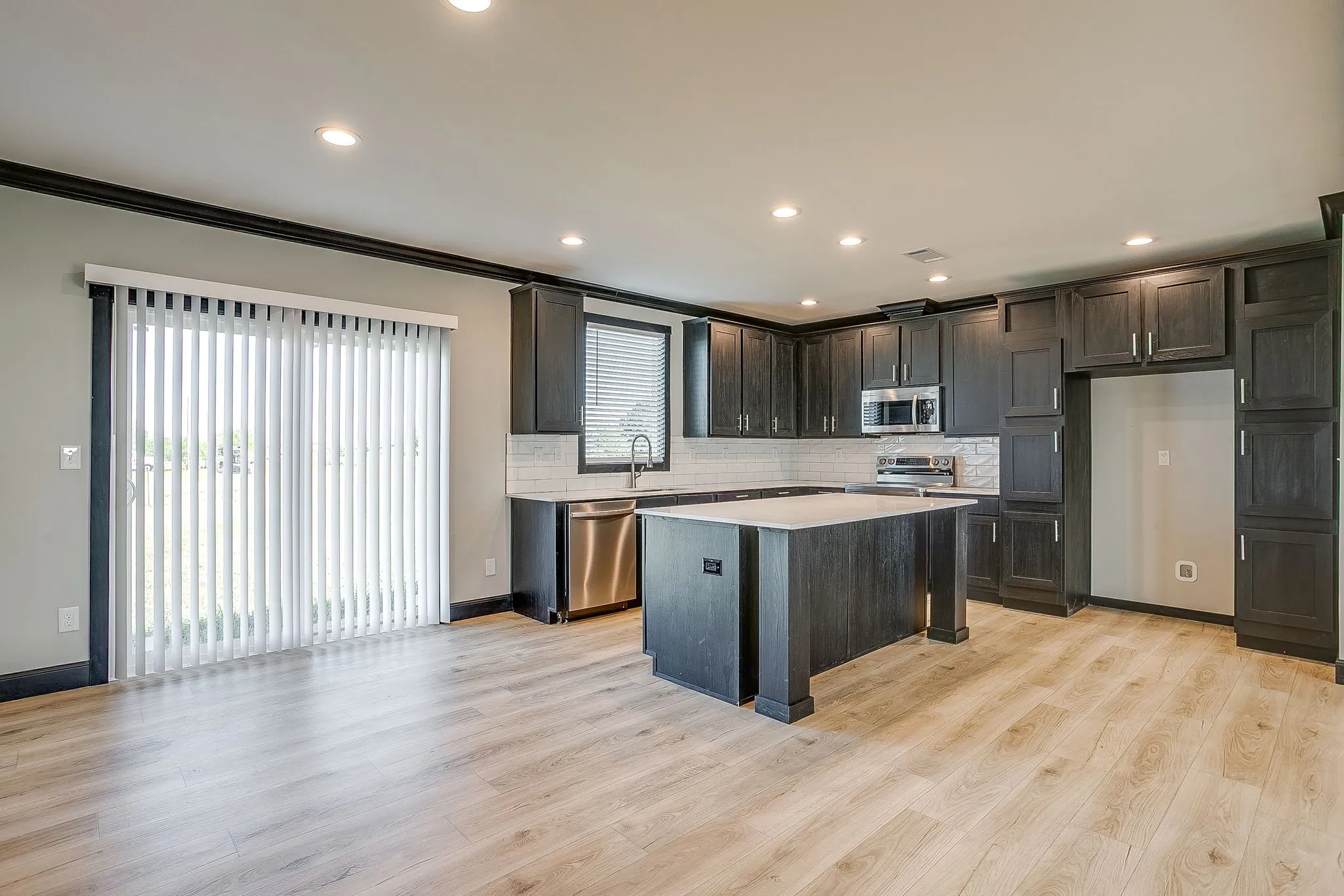 Wood-finish flooring throughout, dark wood-finish cabinetry, light-toned countertops, subway tile backsplash, and stainless steel appliances