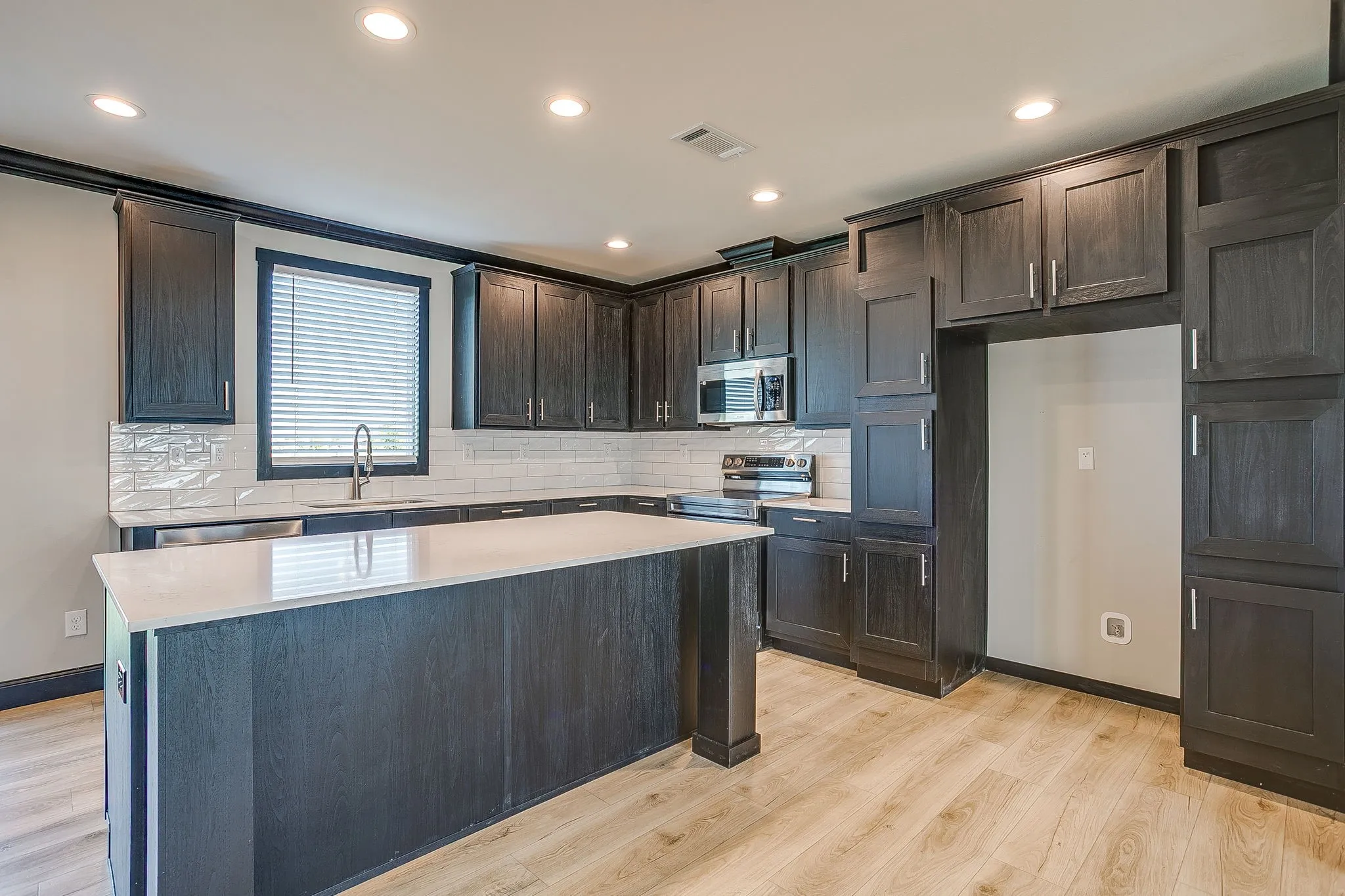 Kitchen featuring wood-finish flooring, dark wood-finish cabinetry, a large center island with a light-toned countertop, subway tile backsplash, and recessed lighting