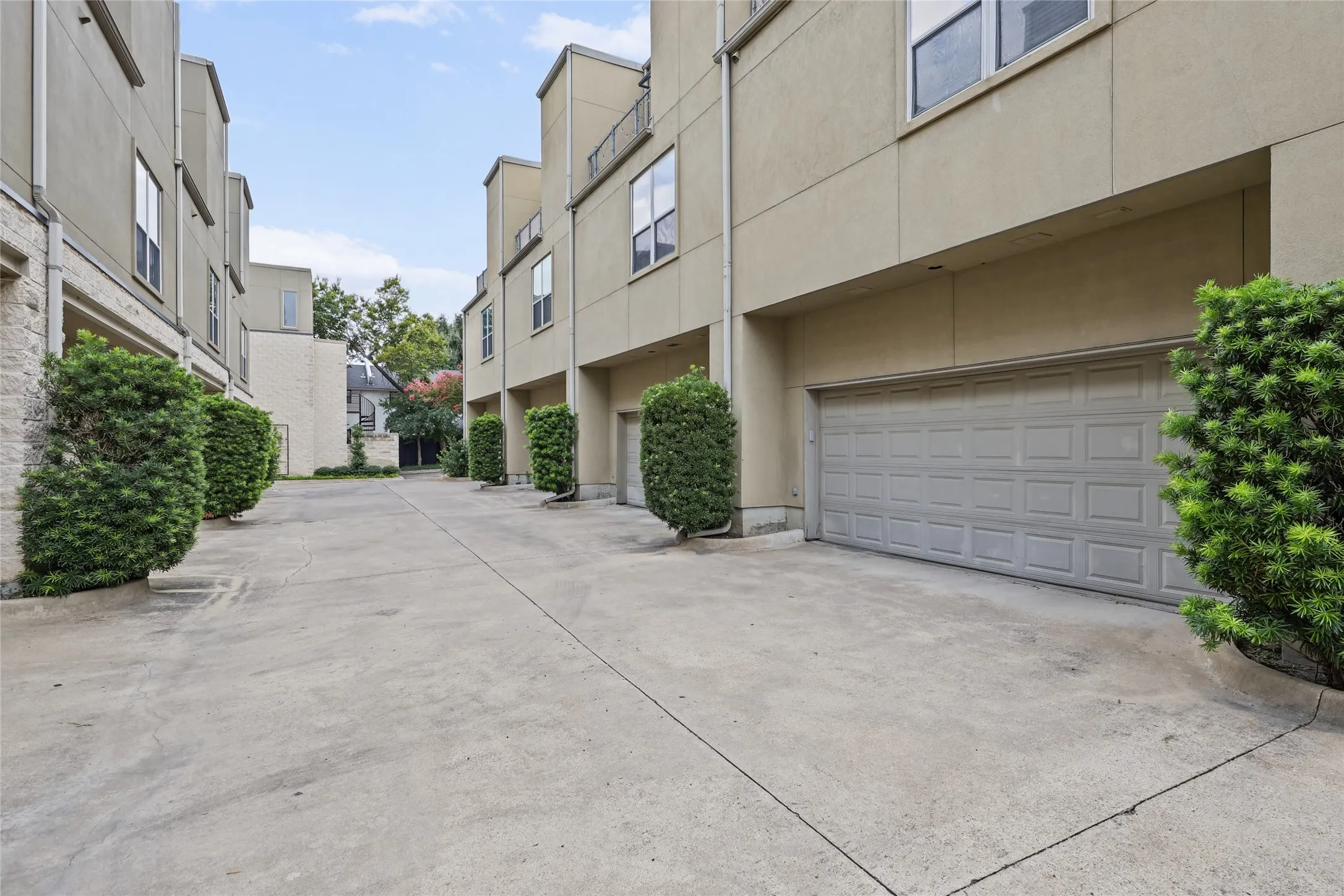 Gated courtyard featuring smooth concrete hardscaping, dual garage doors with modern gray finish, and mature landscaping with sculpted hedges